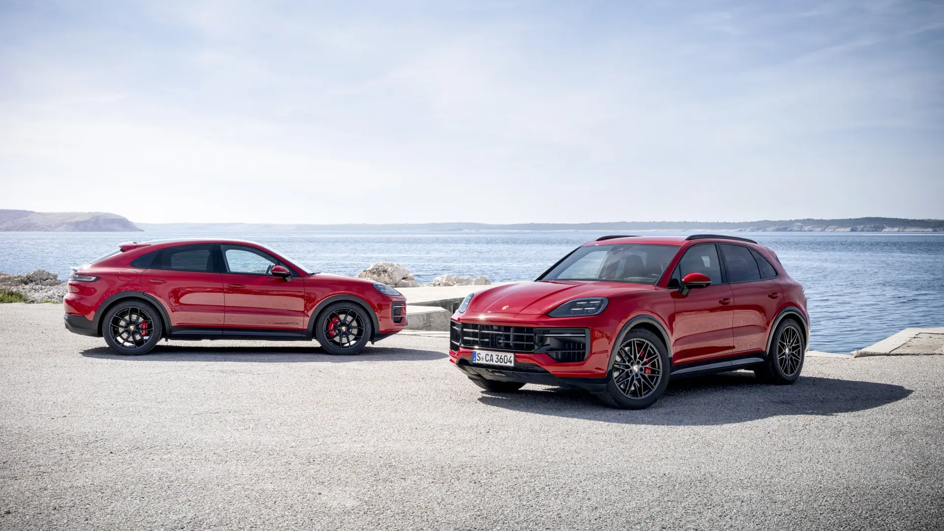 To røde Porsche Cayenne GTS SUV’er parkeret ved en havnepromenade med hav og klipper i baggrunden.
