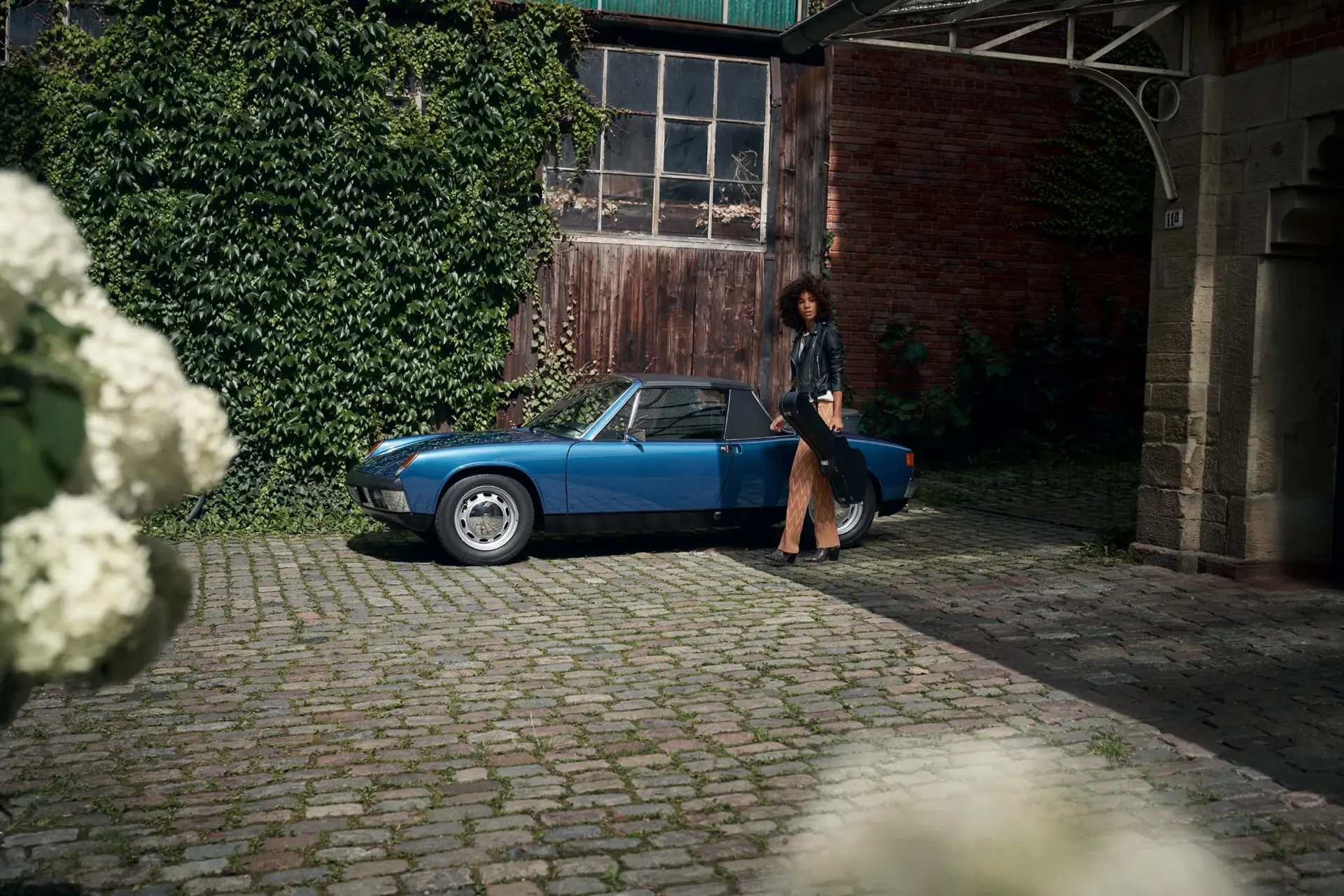 En person står med et instrumentetui ved siden af en blå klassisk Porsche parkeret i en brostensgård foran en mur med klatreplanter.