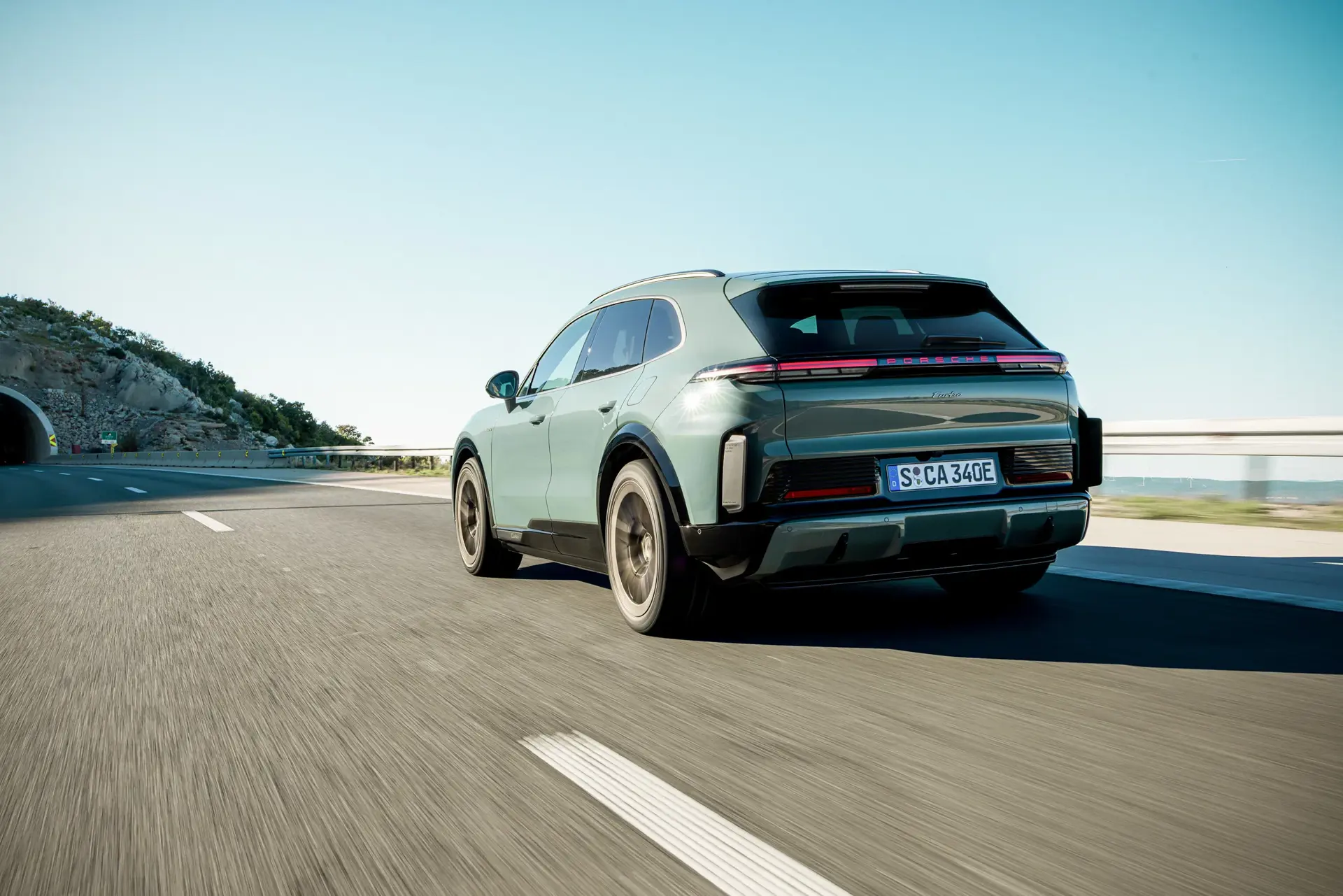 En grøn Porsche SUV ses bagfra, mens den kører på en motorvej i solrigt vejr nær en tunnel.