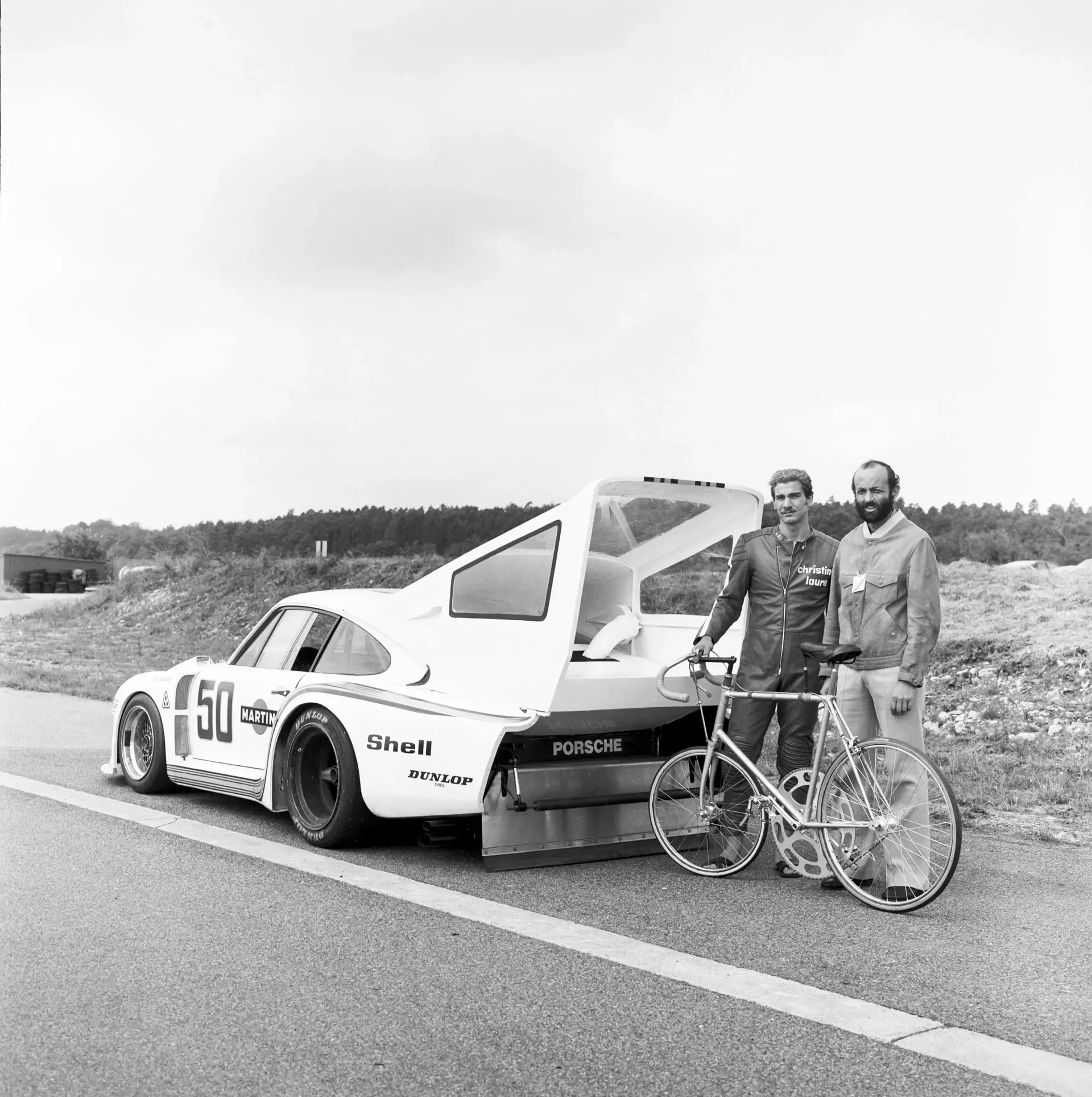 Sort-hvidt foto af to mænd med en racercykel ved siden af en Porsche-racerbil med åben bagklap på en landevej.