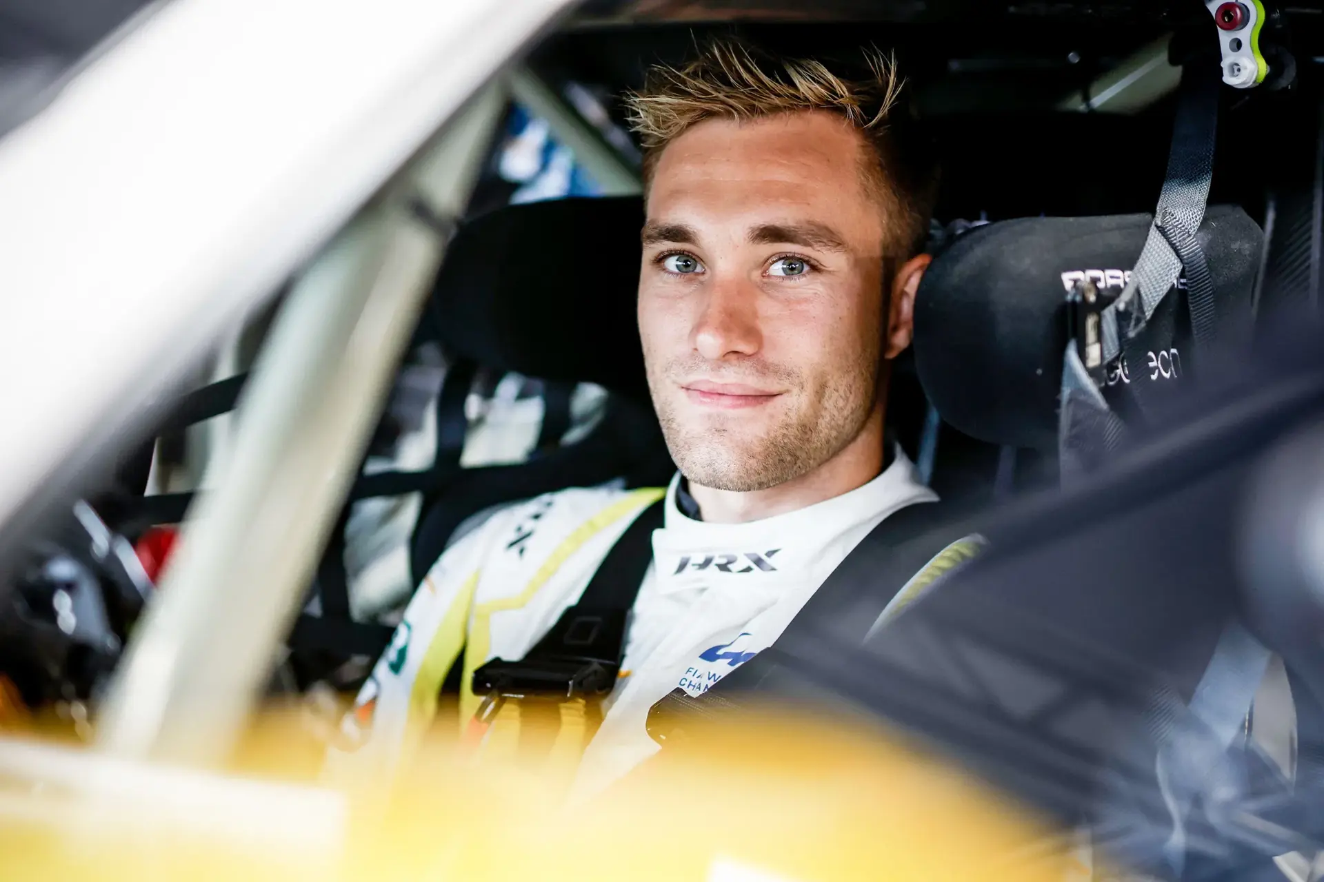 Racerkører i køredragt og sikkerhedsseler sidder klar i førersædet på en racerbane.