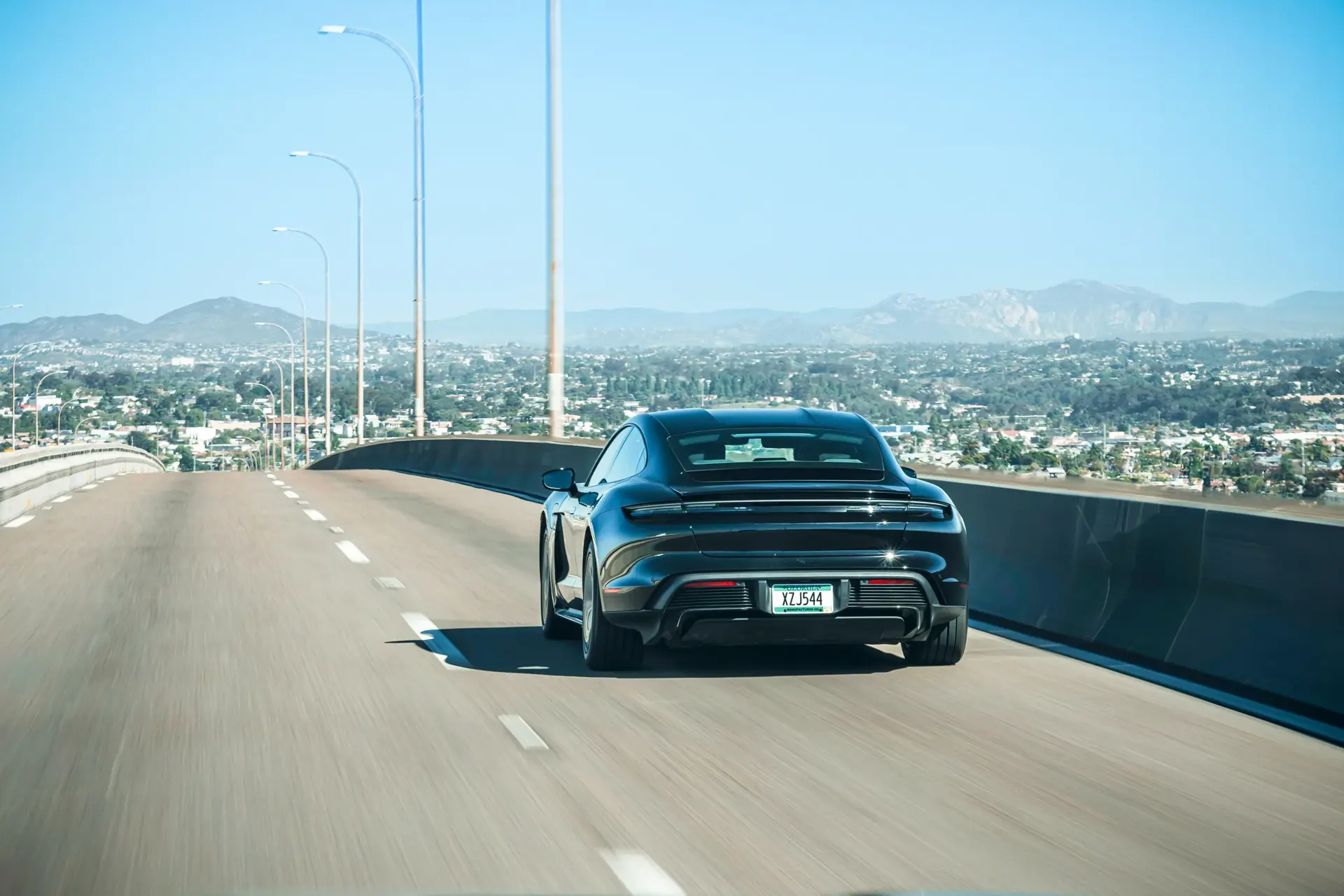 En sort Porsche Taycan kører på en motorvej med udsigt over en by og fjerne bjerge i klart solskin.