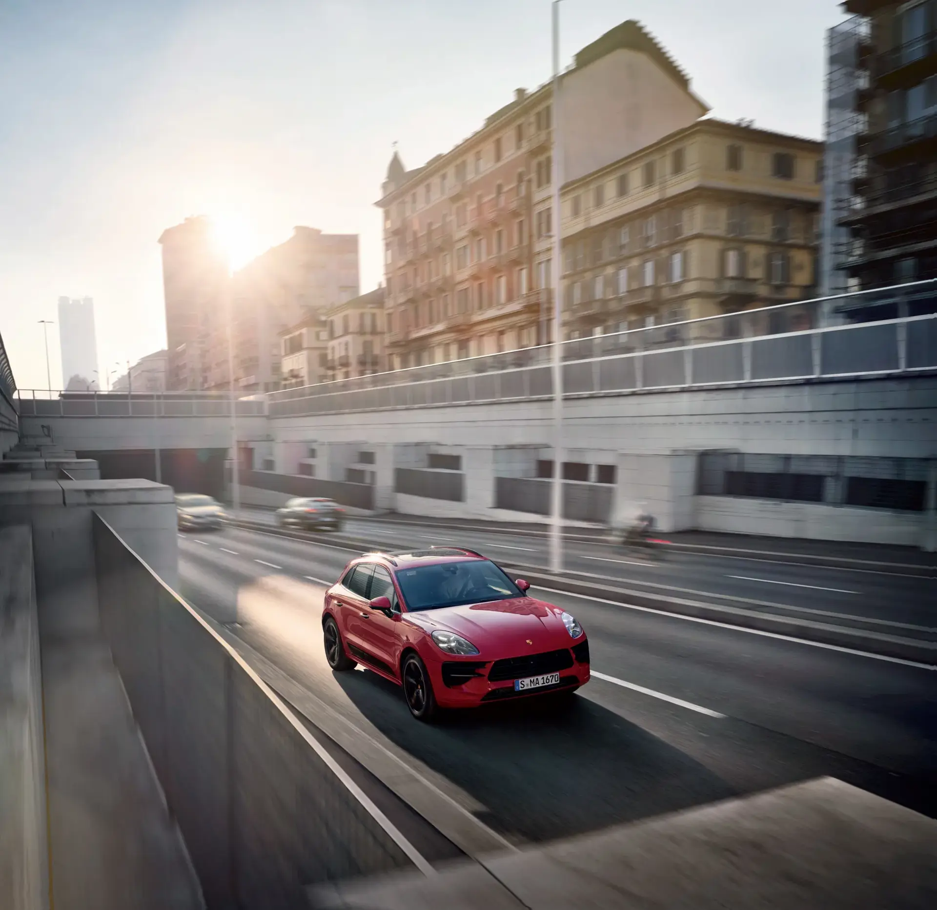 Rød Porsche SUV kører hurtigt på en bymotorvej i morgensol mellem høje bygninger.