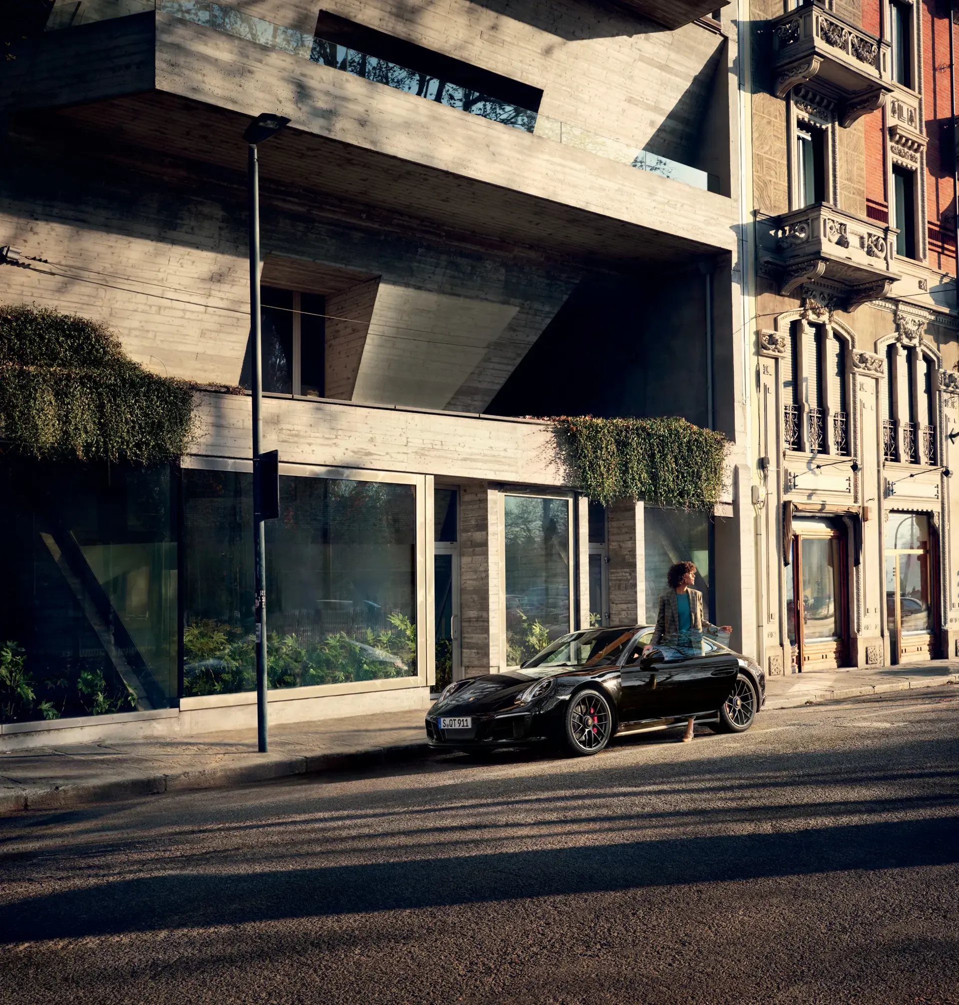 En sort Porsche sportsvogn holder parkeret på en solbeskinnet bygade foran en moderne betonbygning, mens en person står ved siden af bilen.