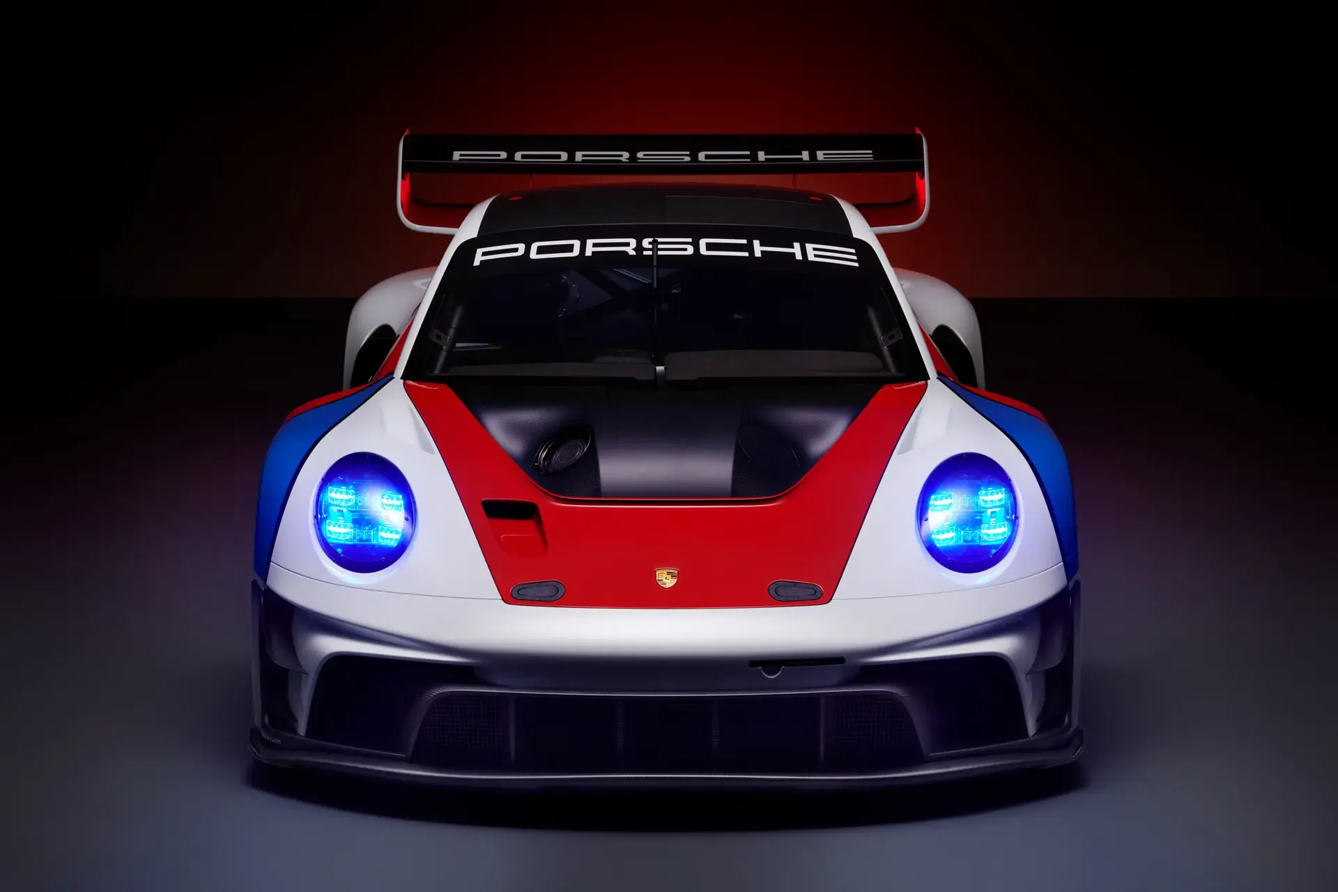 Frontvisning af en hvid, rød og blå Porsche GT3-racerbil med blå forlygter og stor bagvinge mod mørk baggrund.