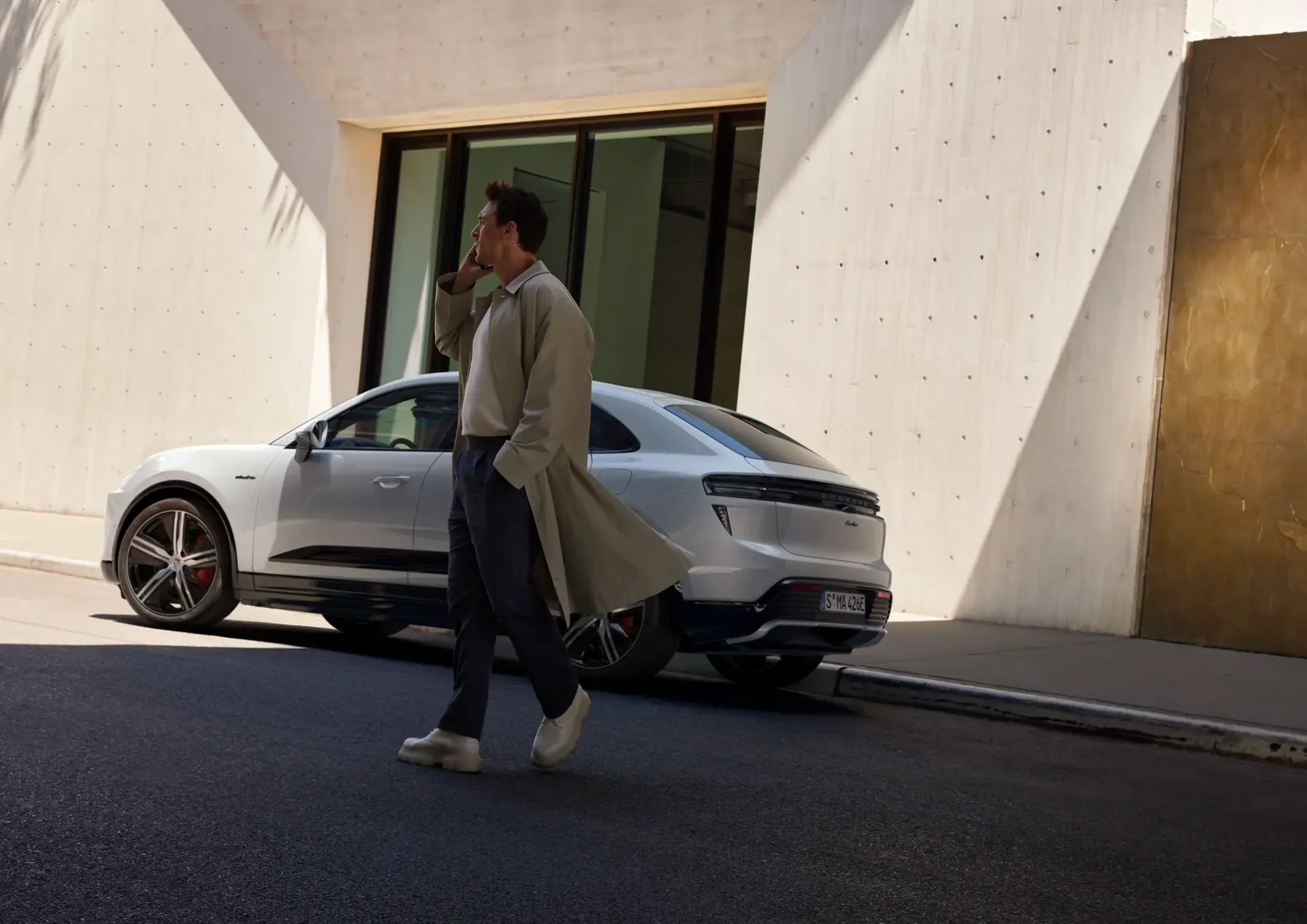 En mand går og taler i telefon foran en hvid Porsche Macan parkeret ved en moderne bygning.