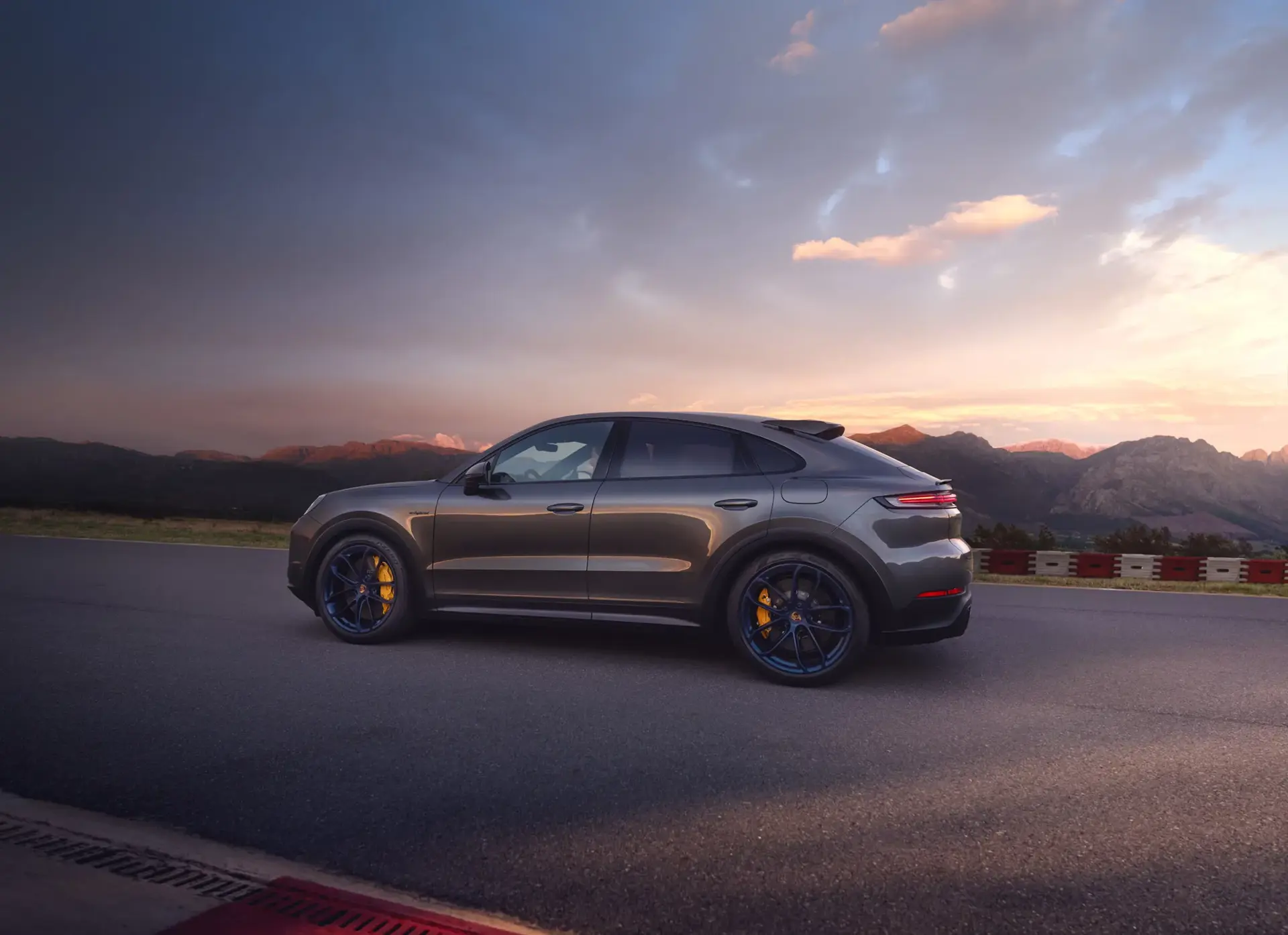 En grå Porsche Cayenne Turbo E-Hybrid Coupé med GT-pakke står på en racerbane foran et bjerglandskab ved solnedgang.