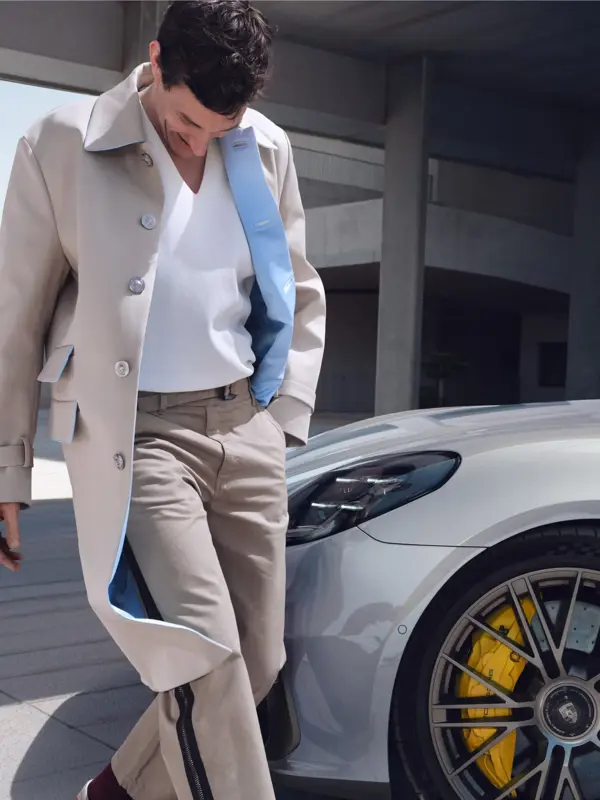 En mand i lys frakke står lænet mod en sølvgrå Porsche Panamera Turbo S E-Hybrid, hvor bilens forhjul med gule bremsekalibre er tydeligt i fokus.