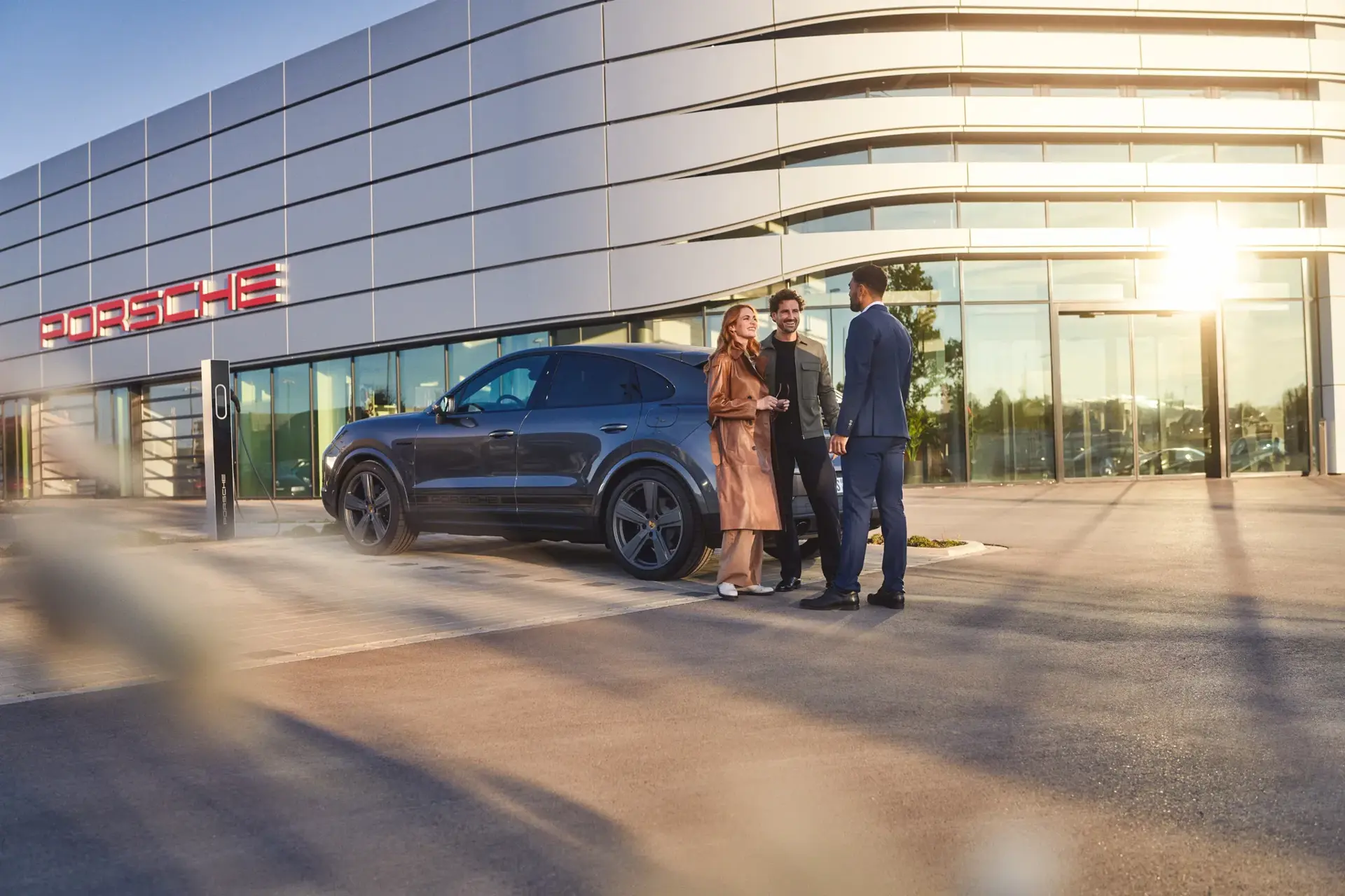 Tre personer står og taler ved en mørkegrå Porsche, der lader ved en ladestander foran et Porsche‑bilhus.