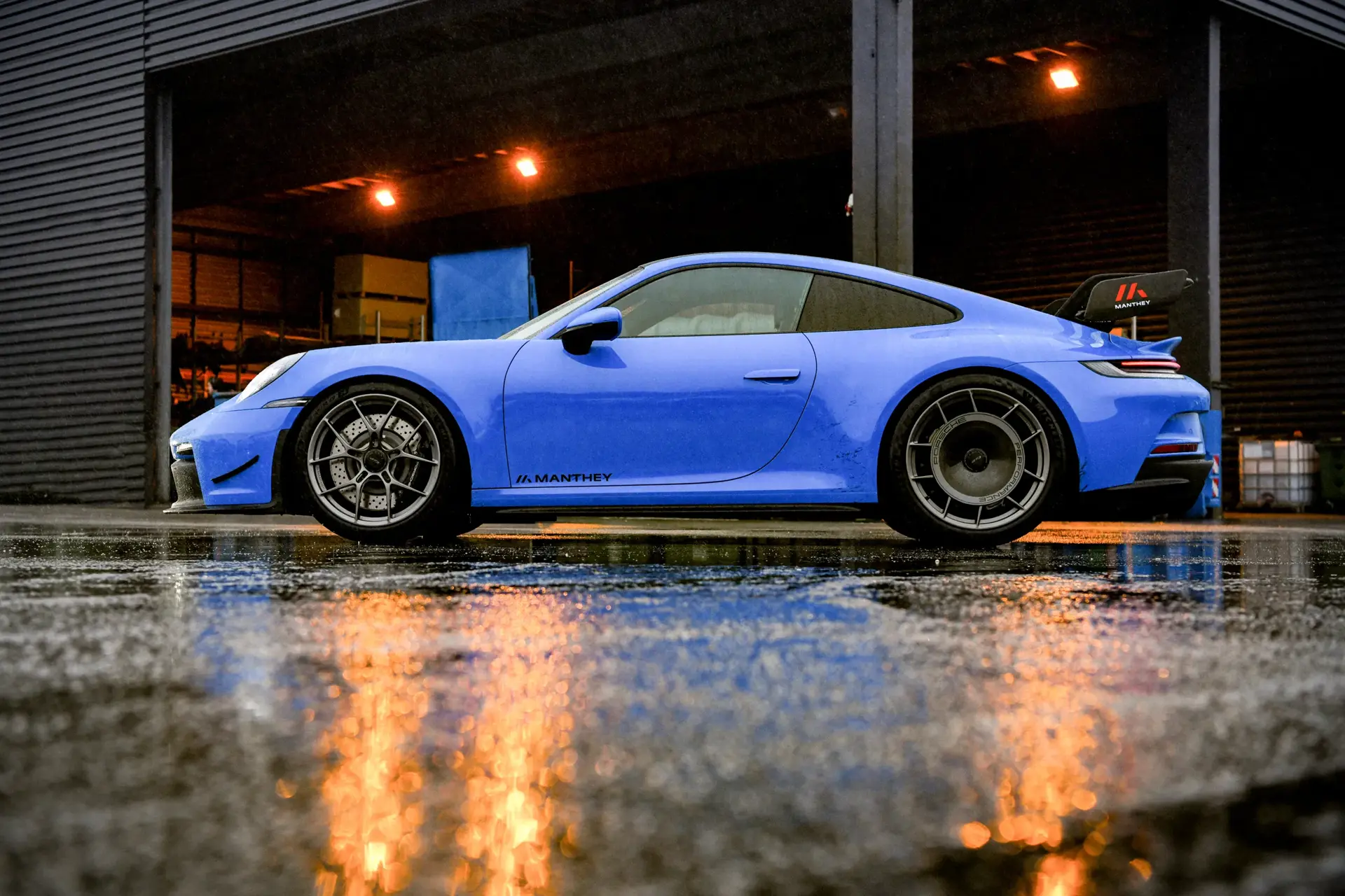 Blå Porsche 911 med Manthey Performance Kit set fra siden foran en åben garage på en regnvåd plads.