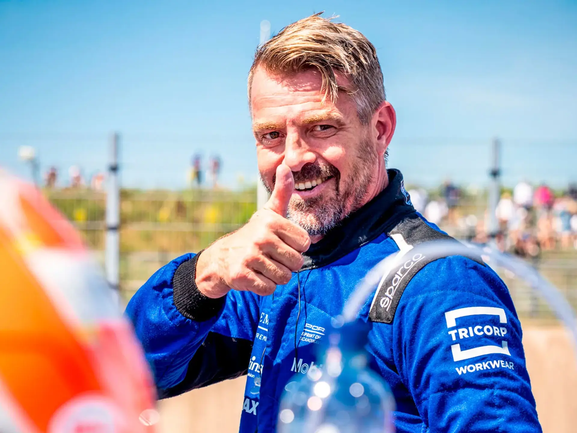 Racerkører i blå køredragt giver thumbs up i pitområdet på en solrig racerbane.