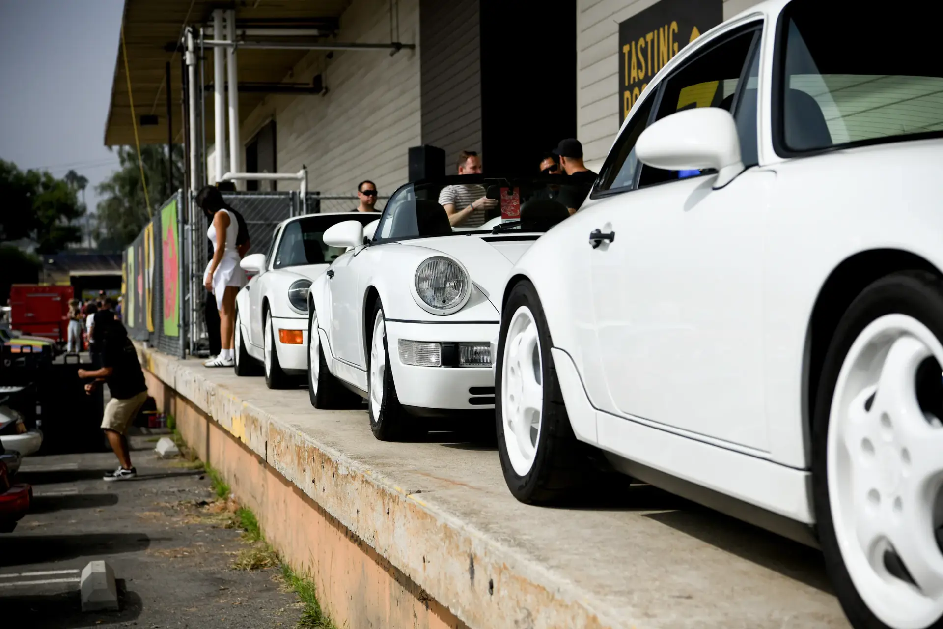 Flere hvide klassiske Porsche 911 står parkeret på en rampe ved et biltræf, mens mennesker samles omkring dem.