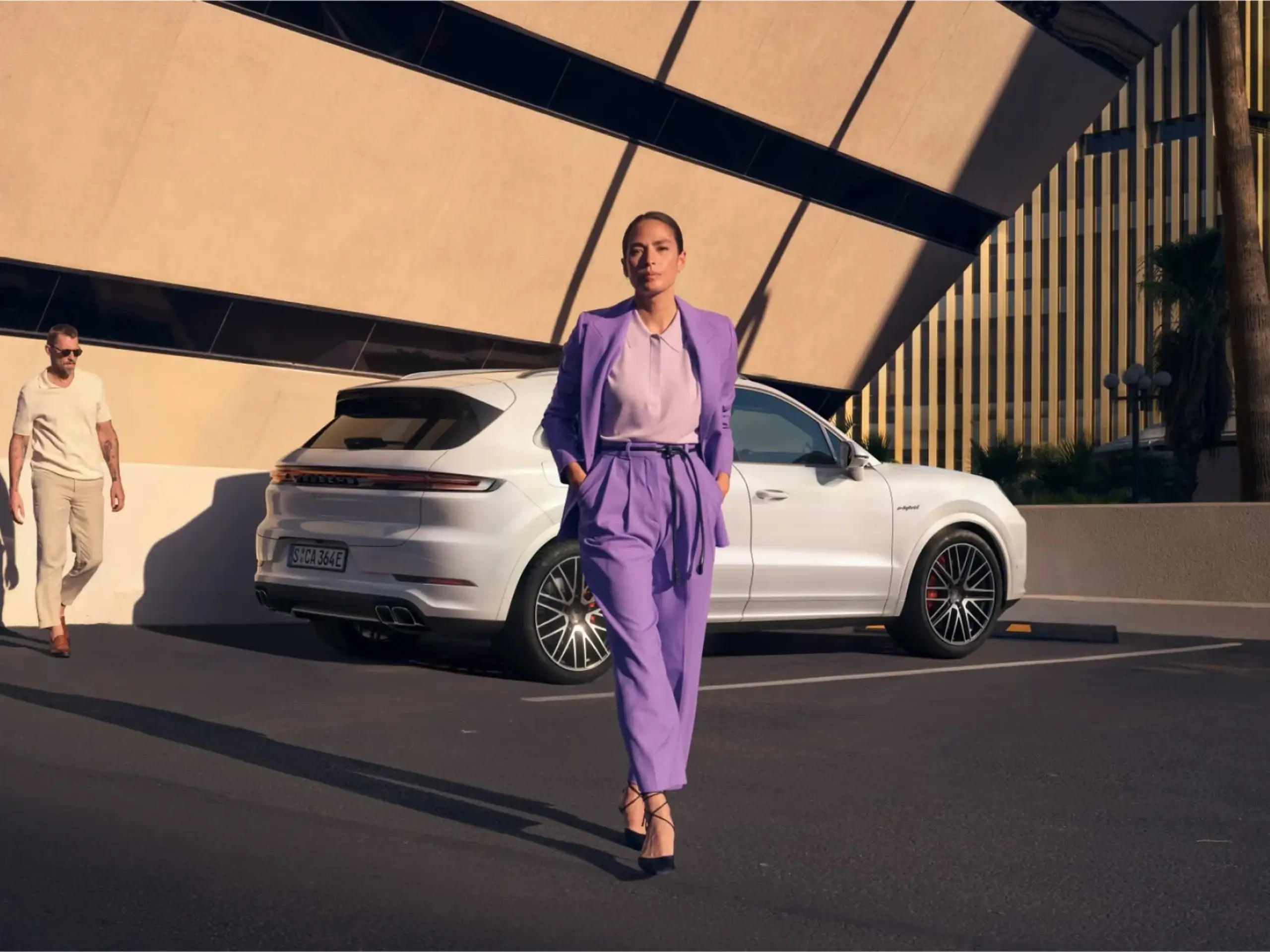 En kvinde i lilla jakkesæt går foran en hvid Porsche Cayenne Turbo E-Hybrid parkeret ved en moderne bygning i sollys.