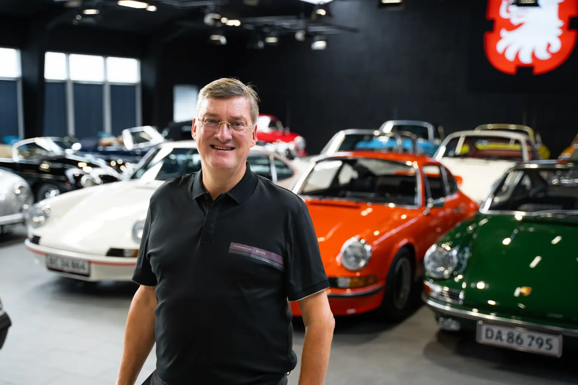 Mand i sort polo står smilende foran en række klassiske Porsche-sportsvogne i et udstillingslokale.
