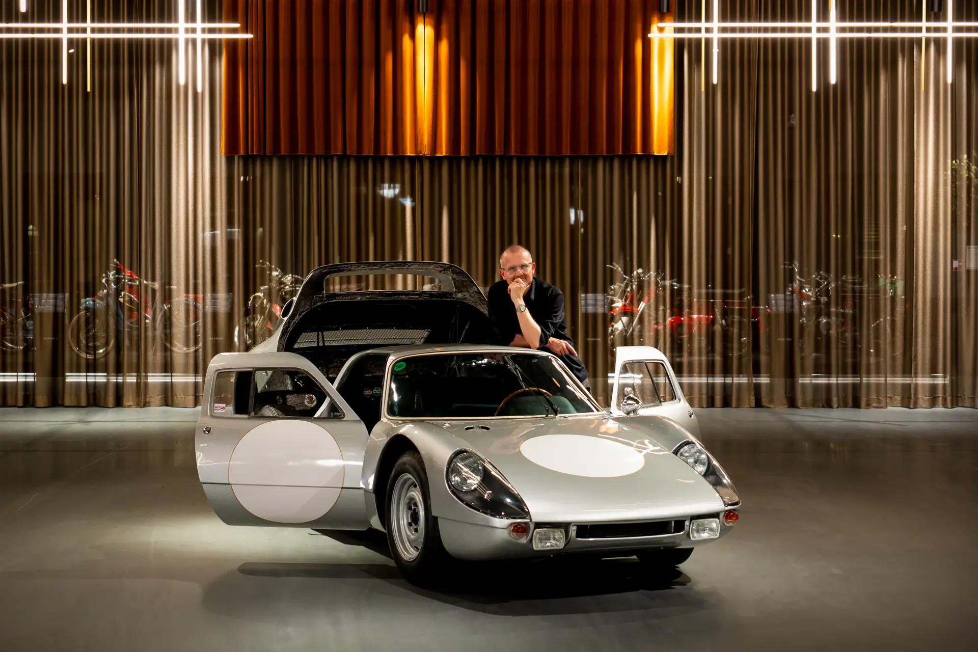 Sølvfarvet klassisk Porsche sportsvogn med åbne døre står udstillet indendørs, mens en person læner sig over bilen.