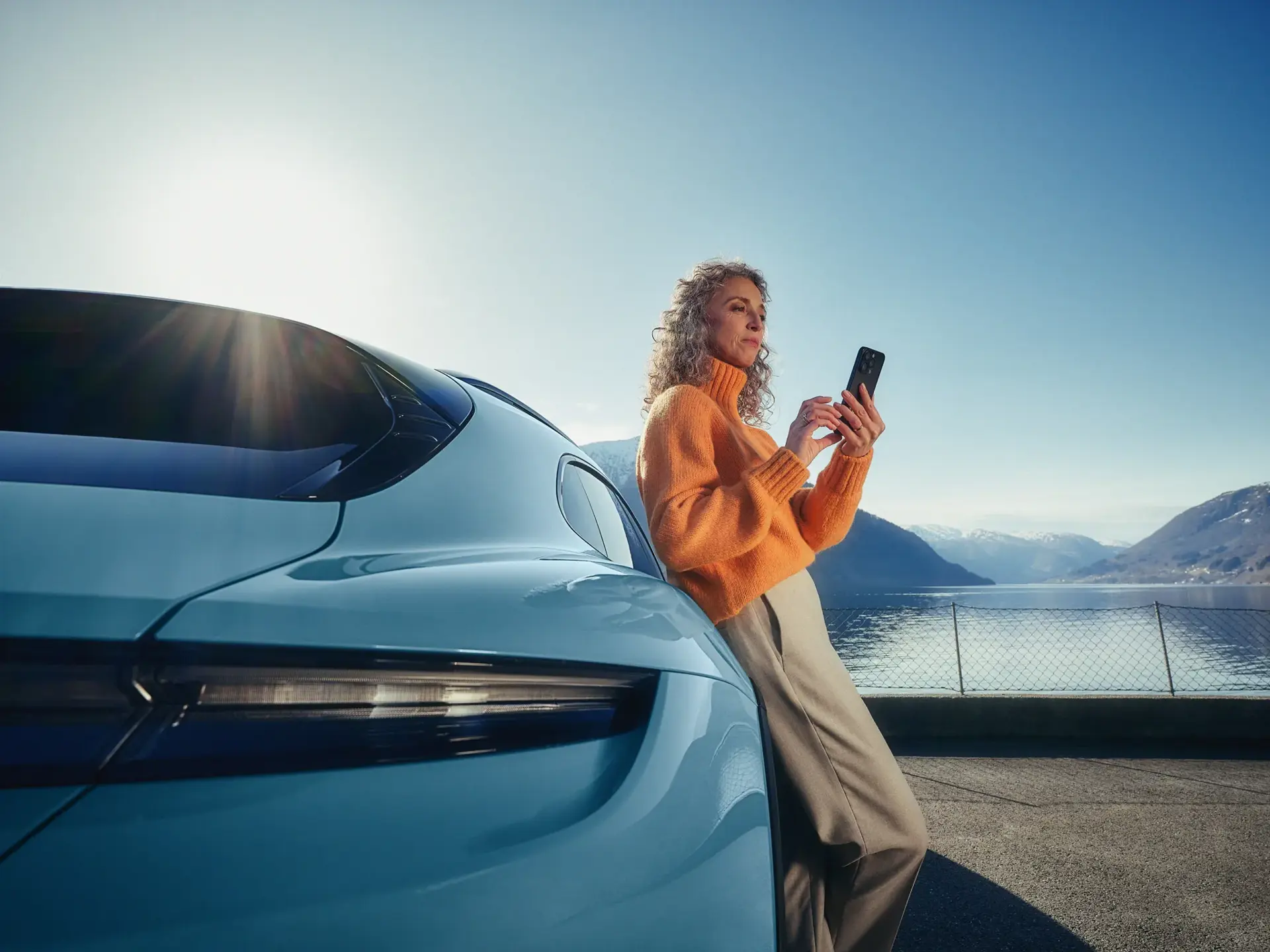 Kvinde læner sig op ad en parkeret bil og bruger sin smartphone ved en sø med bjerge i baggrunden.