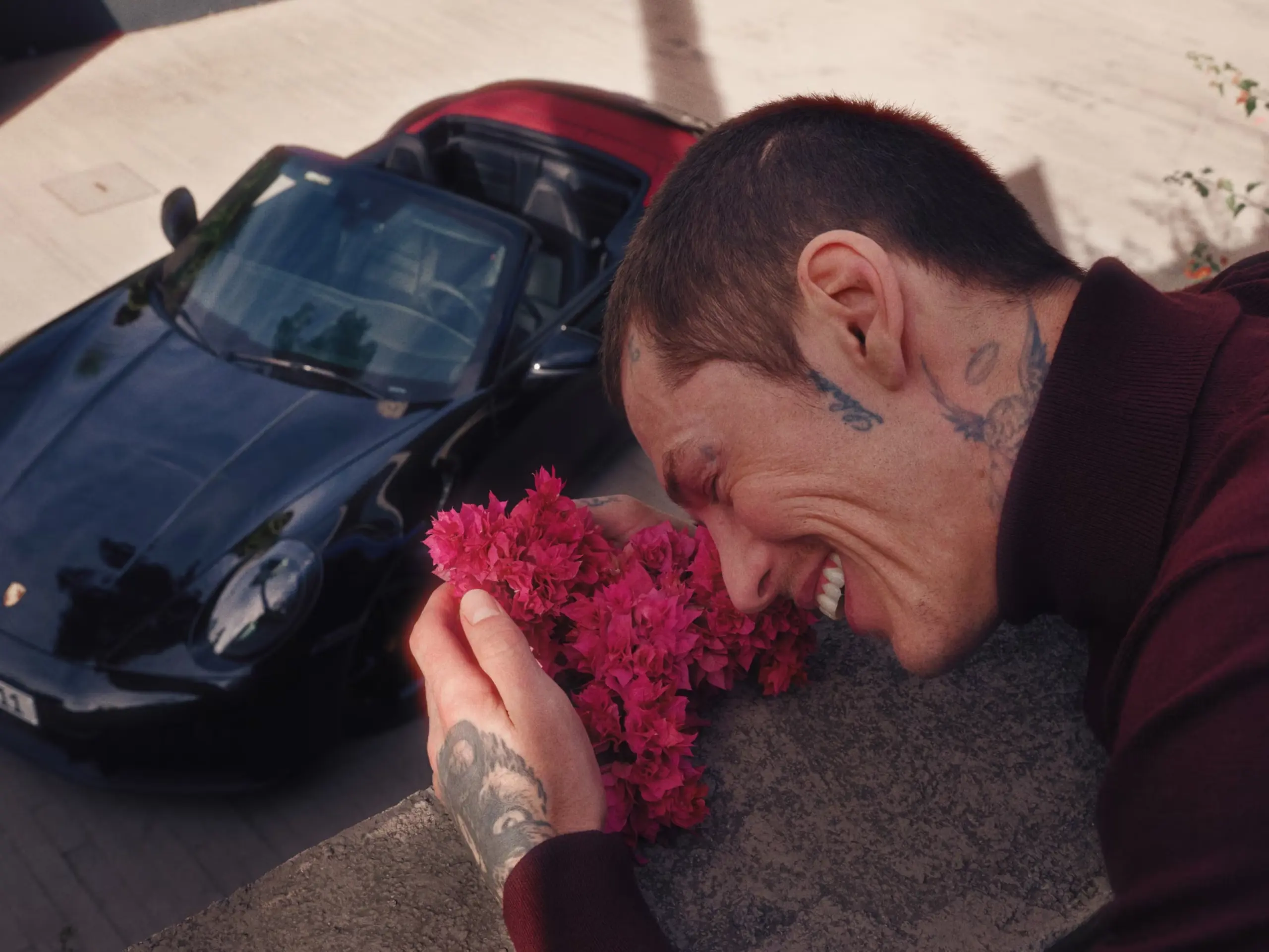 Smilende person med tatoveringer dufter til pink blomster, mens en sort Porsche 911 Cabriolet holder parkeret nedenfor.