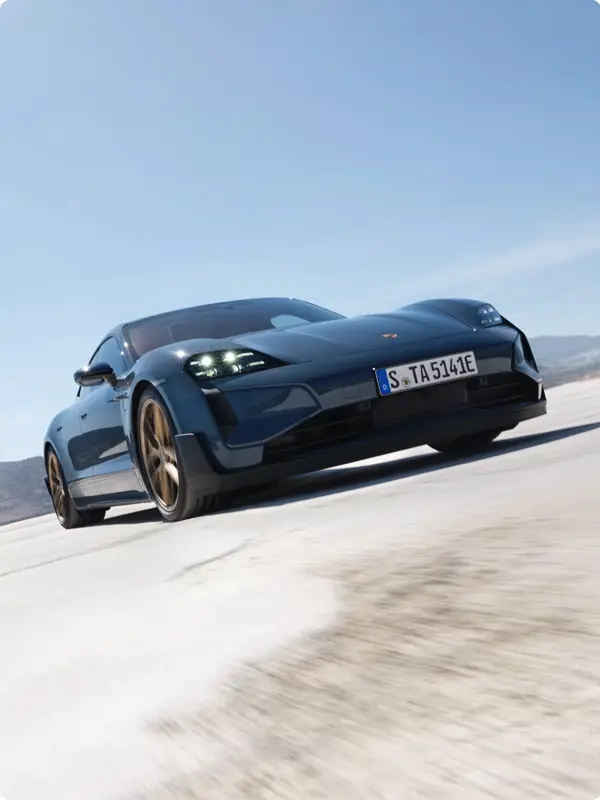 Mørkeblå sportsvogn, formentlig en Porsche Taycan, ses i fart på en åben vej under blå himmel.