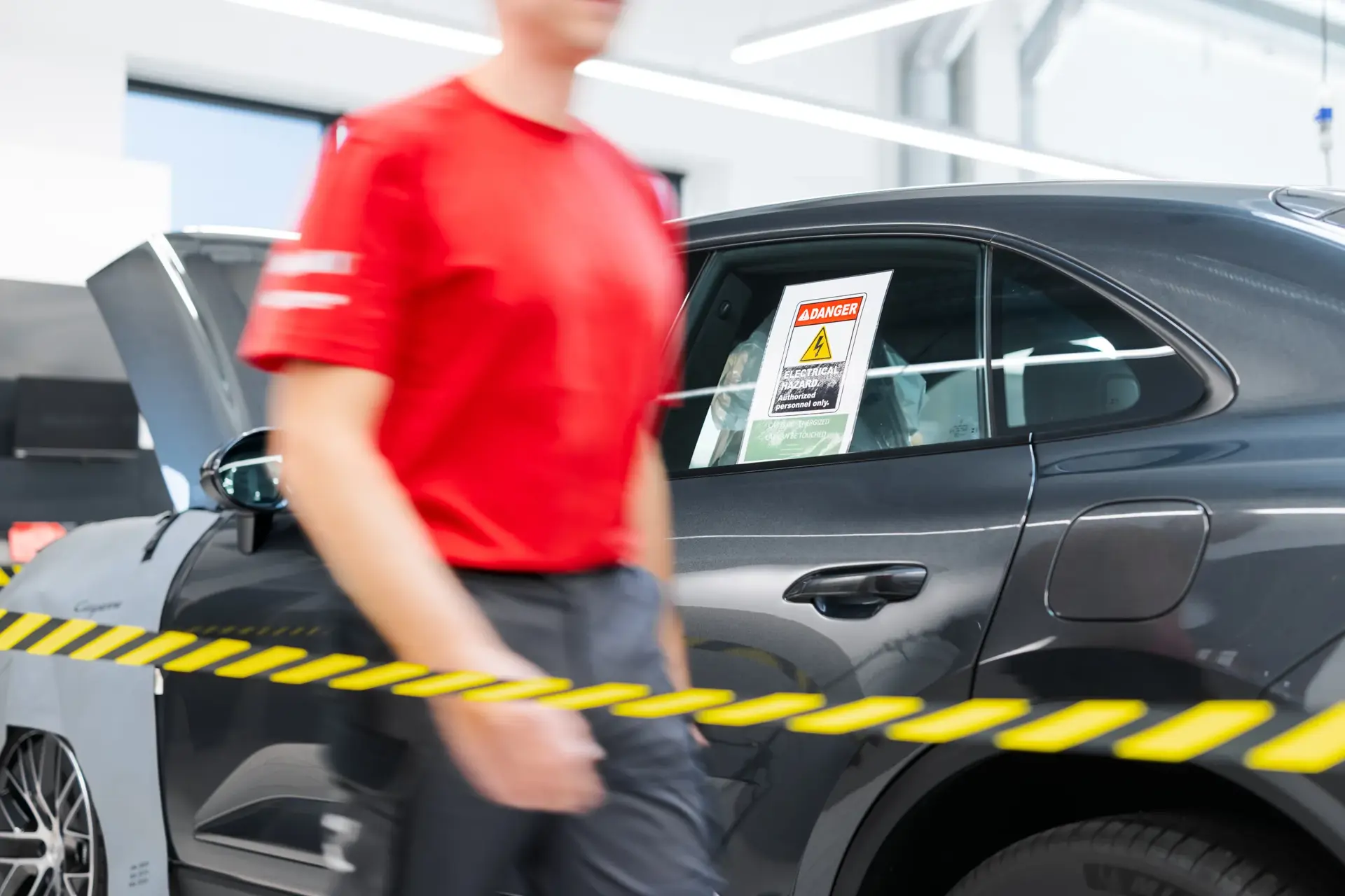 Porsche-bil på værksted med advarselsskilt i ruden og en mekaniker i rød trøje bag afspærringstape.