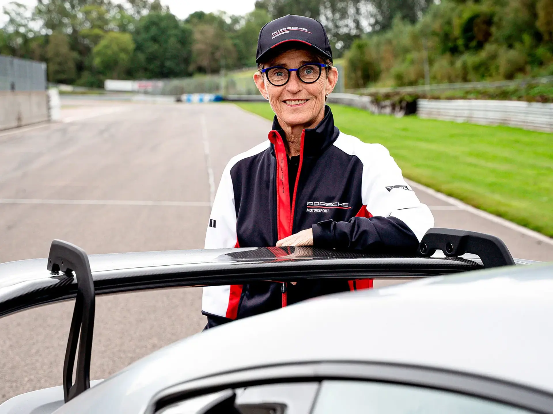 Person i Porsche Motorsport-tøj står ved en sølvfarvet sportsvogn på en racerbane.