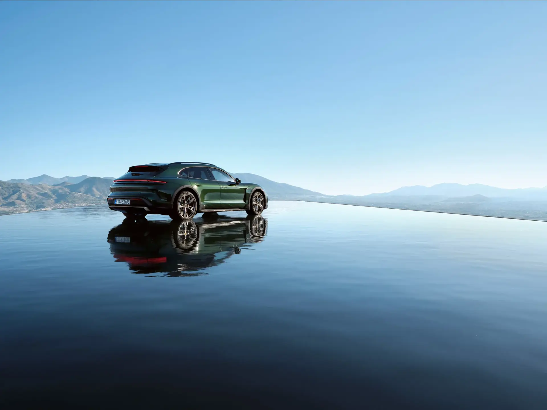 Grøn Porsche Taycan Cross Turismo parkeret ved en spejlblank vandflade med bjerge i baggrunden under en klar blå himmel.