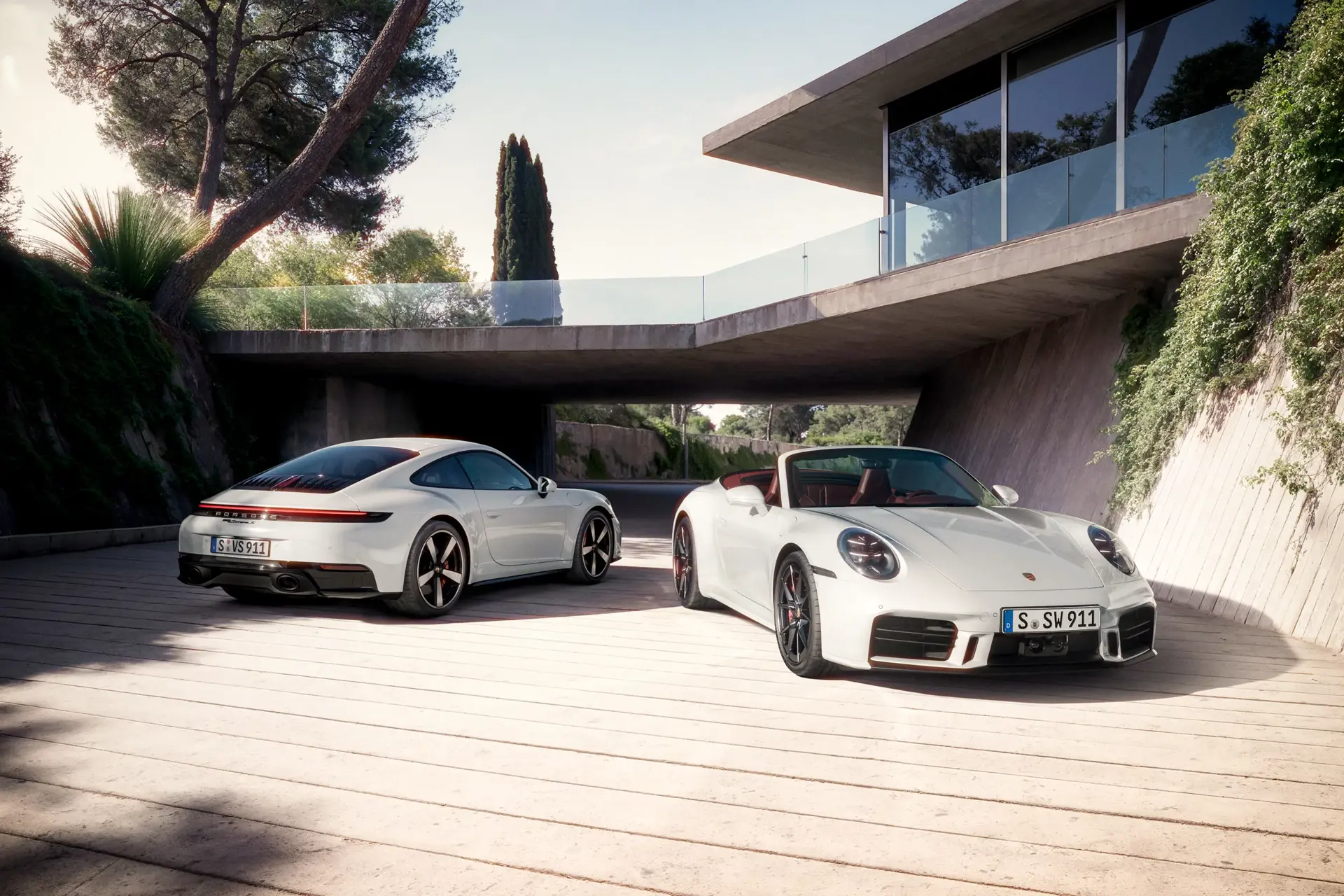 To hvide Porsche 911 Carrera S, en coupé og en cabriolet, holder parkeret foran en moderne villa i sollys.