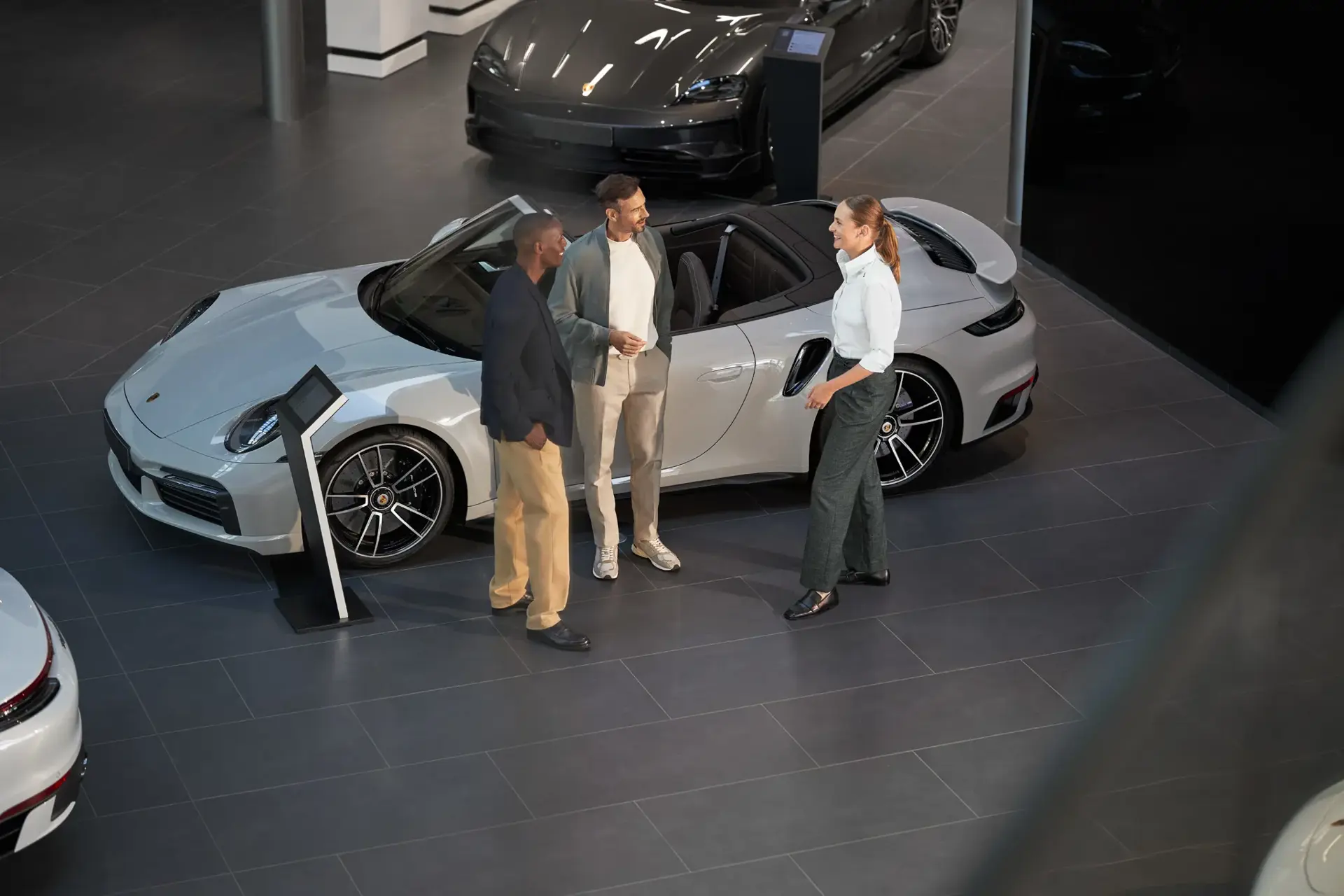 Tre personer står og taler ved en sølvfarvet Porsche-sportsvogn i et moderne bilshowroom.