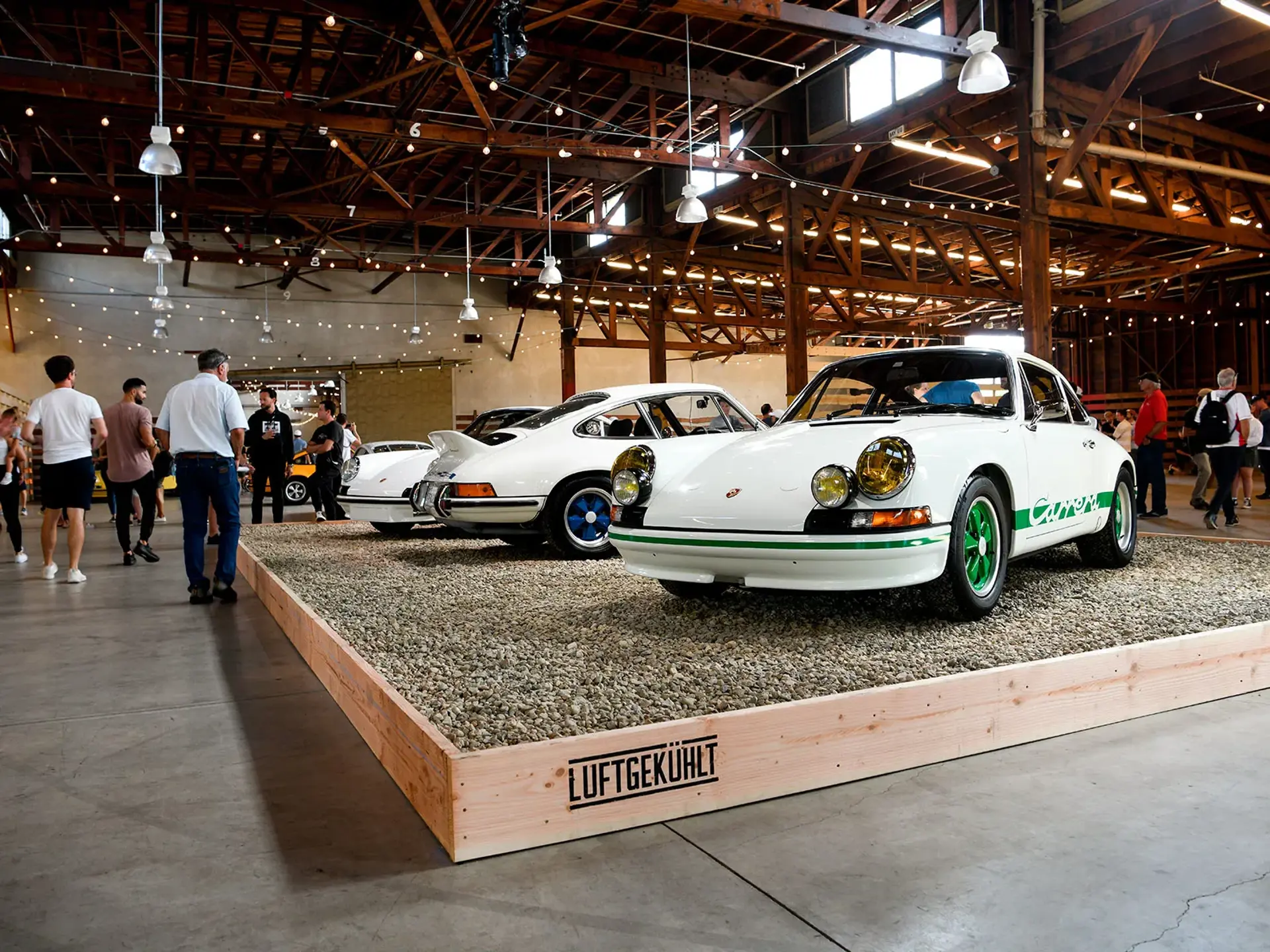 Klassiske hvide Porsche sportsvogne udstilles på en forhøjet stand indendørs ved et Luftgekühlt-arrangement med besøgende omkring.