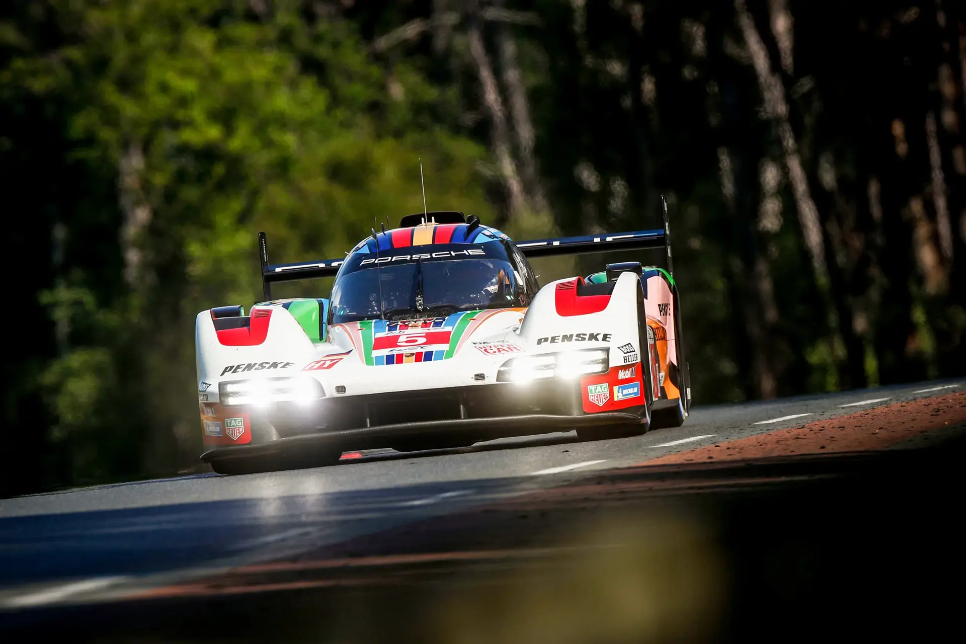 En Porsche-racerbil i farverigt Penske-design kører i høj fart på en racerbane omgivet af træer.