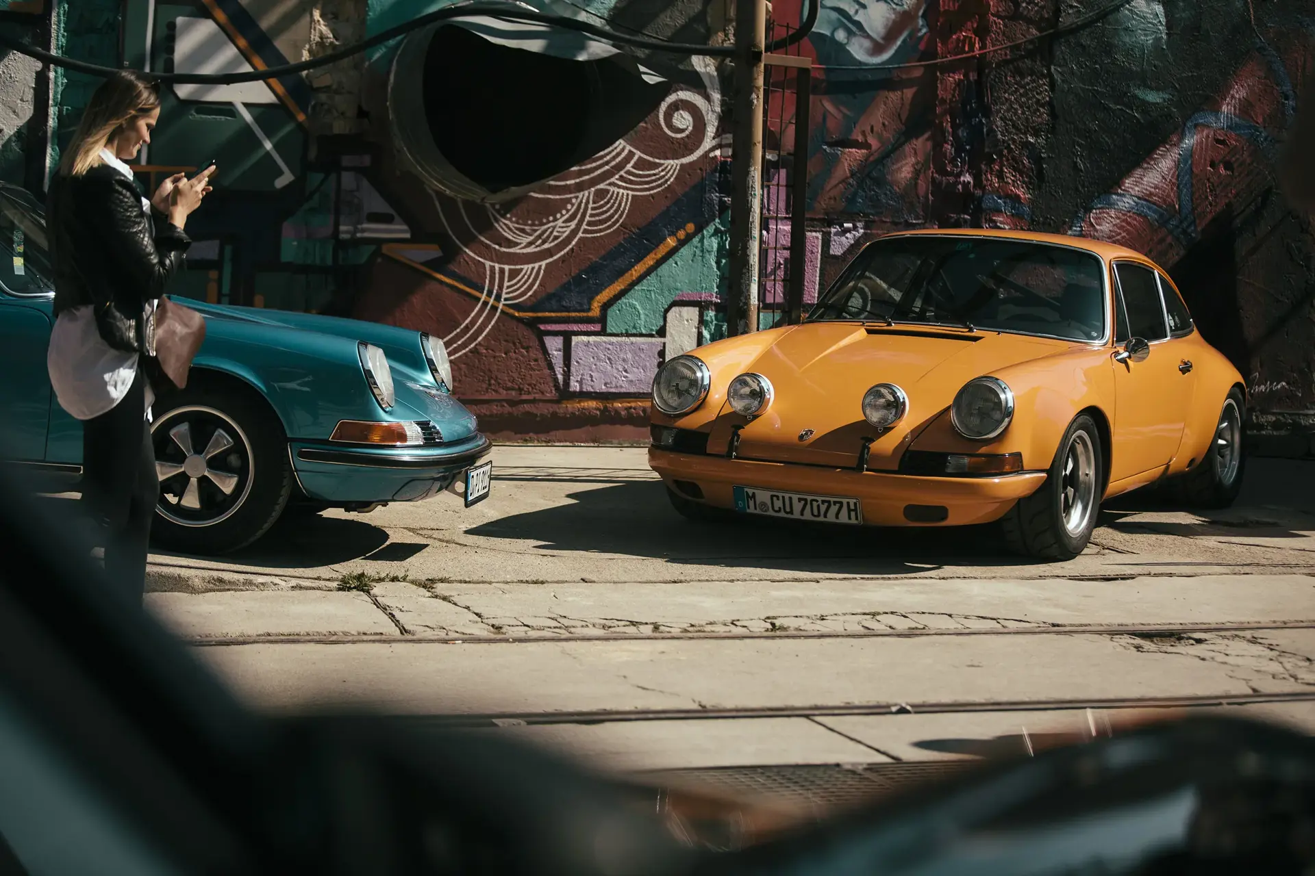 To klassiske sportsvogne, en turkis og en orange, holder parkeret foran en farverig graffitivæg, mens en person går forbi.
