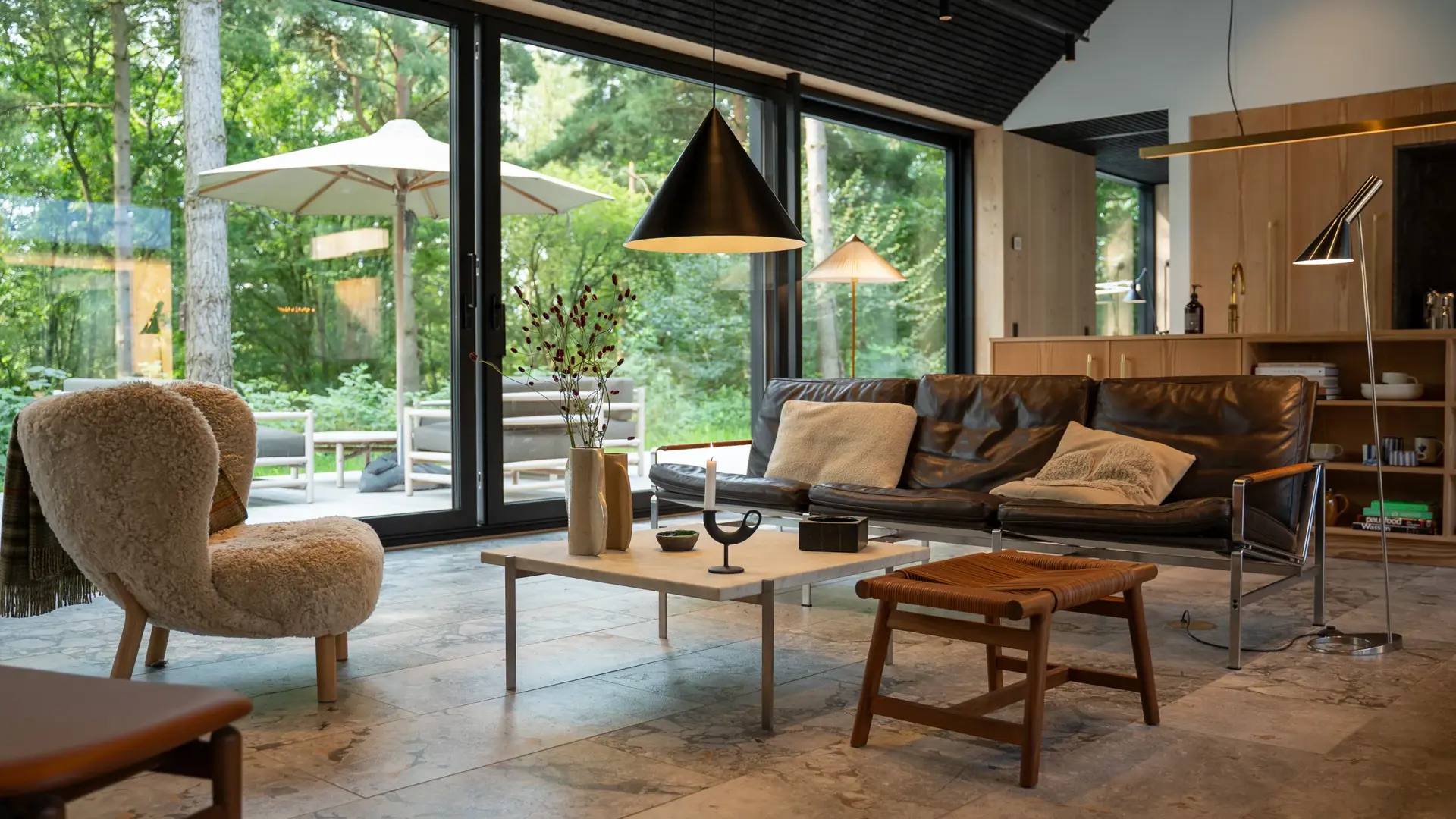 Lys og moderne stue med store glaspartier mod en grøn have, lædersofa, loungestol og stilfuld belysning.