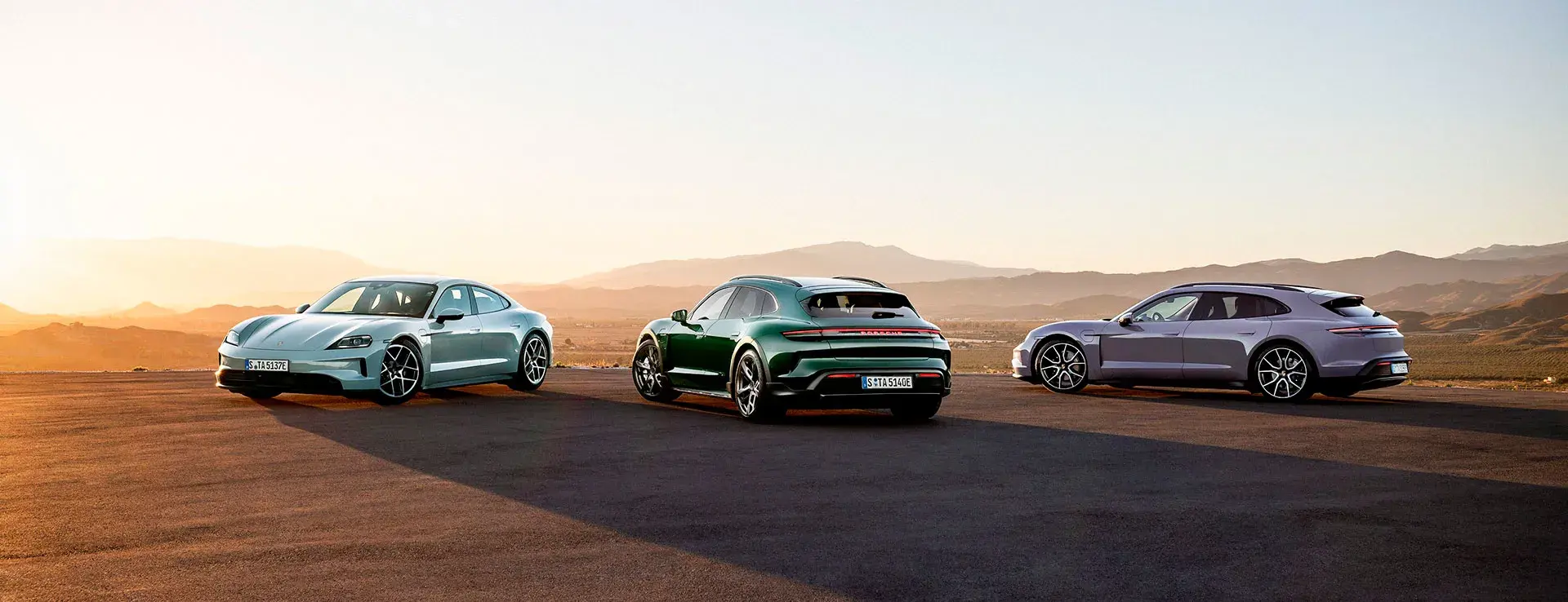 Tre Porsche Taycan-biler står parkeret på en åben vej med bjerglandskab i baggrunden ved solnedgang.
