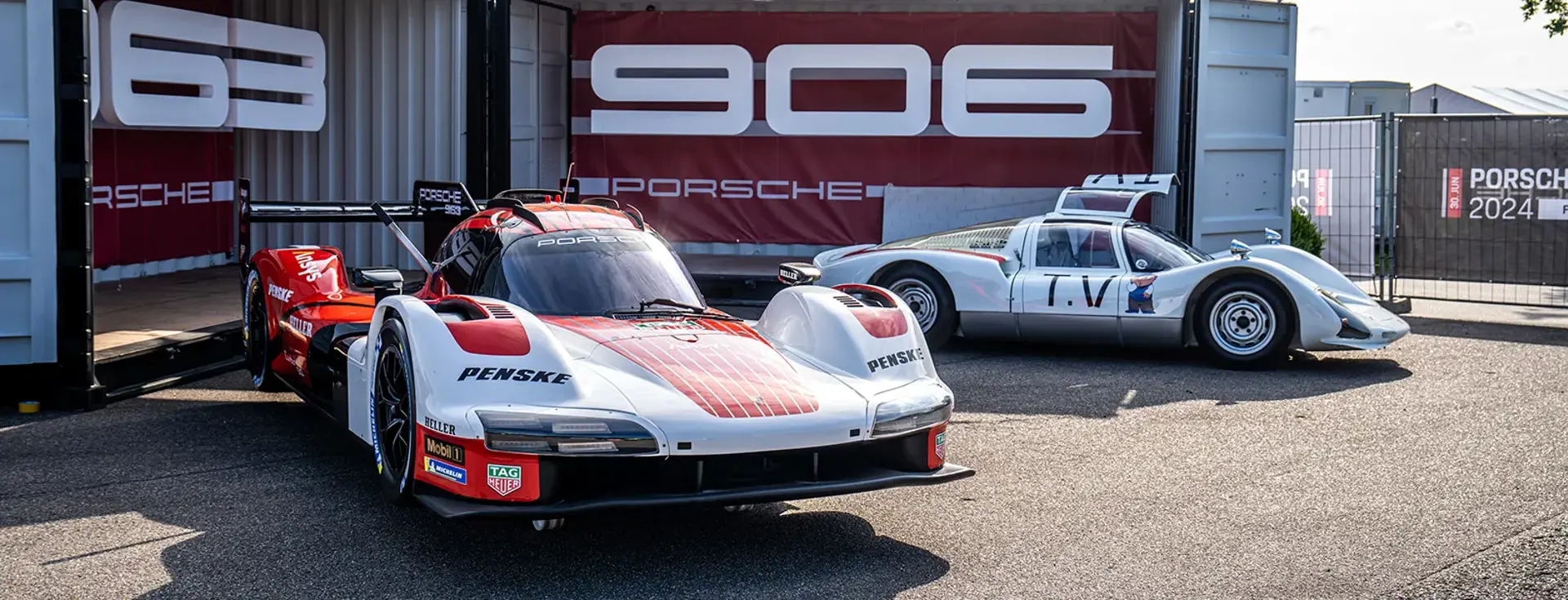 To Porsche-racerbiler, en moderne og en klassisk, står udstillet foran en container med stort Porsche-logo ved en Porsche Festival.