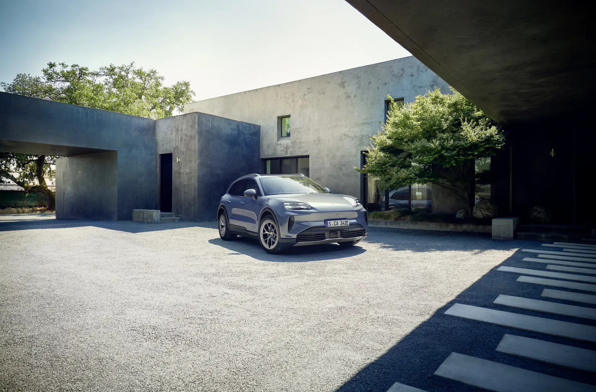 En grå Porsche SUV holder parkeret i gården foran en moderne, minimalistisk betonvilla med grønne træer omkring.