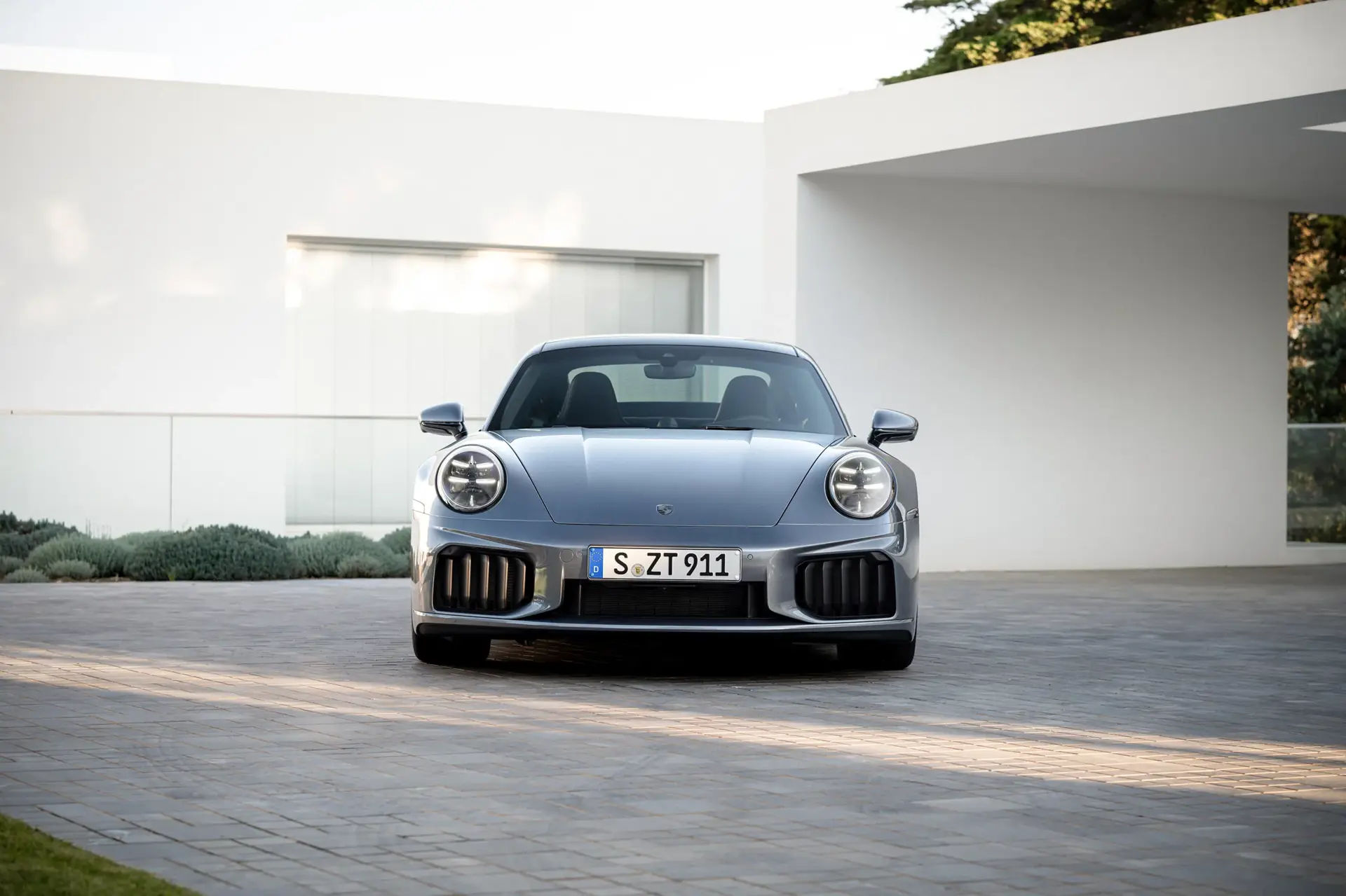 Sølvgrå Porsche 911 set forfra parkeret foran en moderne, hvid villa.