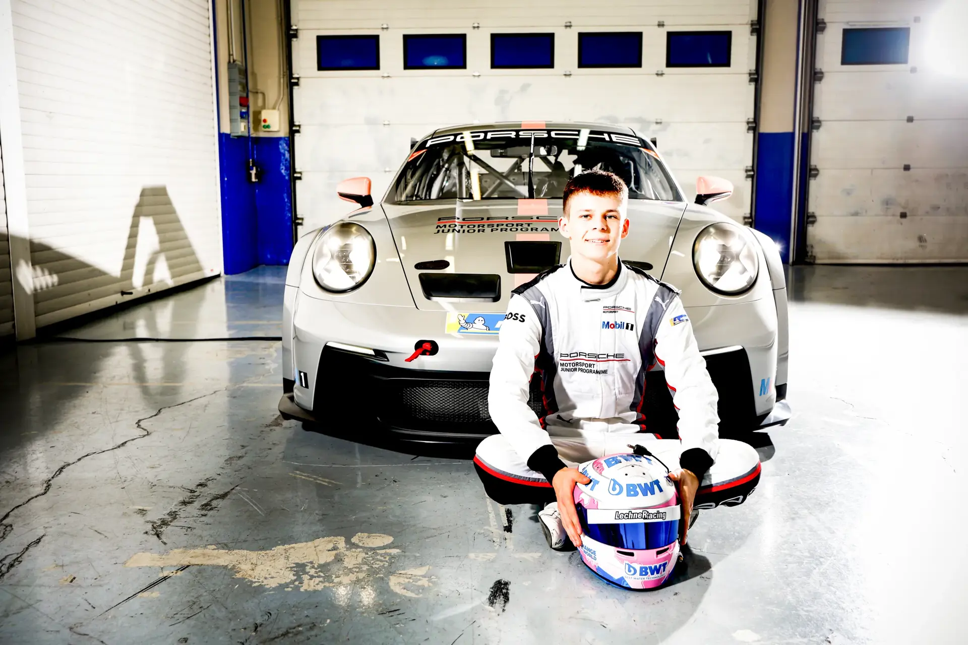 Ung racerkører i Porsche-kørerdragt sidder foran en sølvfarvet Porsche-racerbil i en garage og holder sin blå-rosa hjelm.