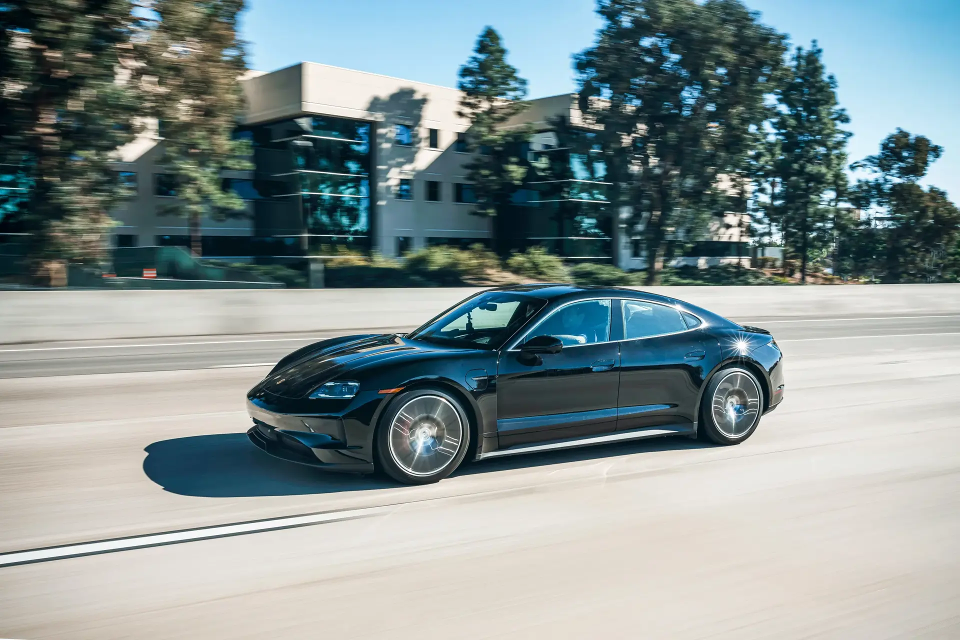 Sort Porsche Taycan elbil i fart på en motorvej med kontorbygninger og træer i baggrunden.