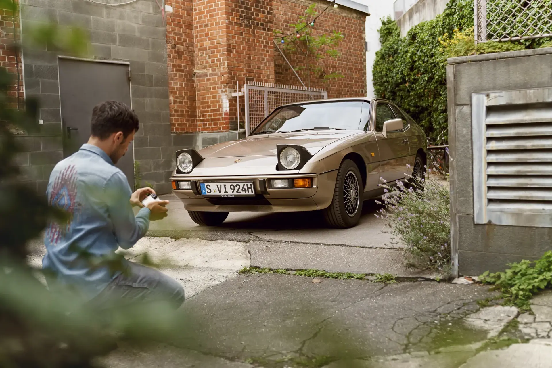 En mand fotograferer en klassisk gylden Porsche sportsvogn i en smal gård mellem murstensbygninger.