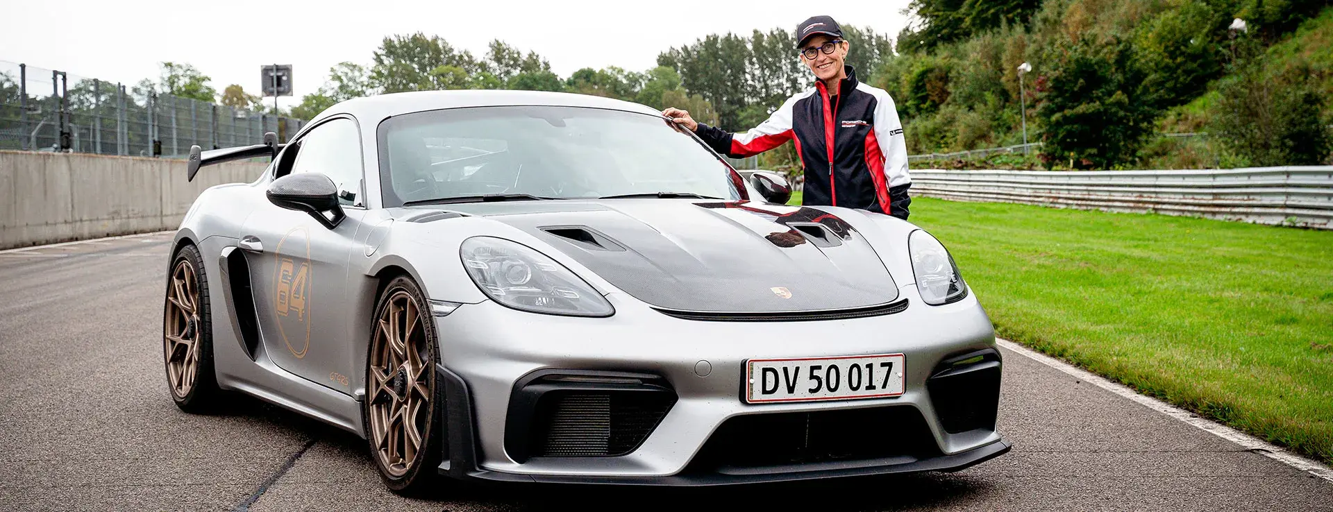 En person i racertøj står ved siden af en sølvgrå Porsche sportsvogn parkeret på en racerbane.