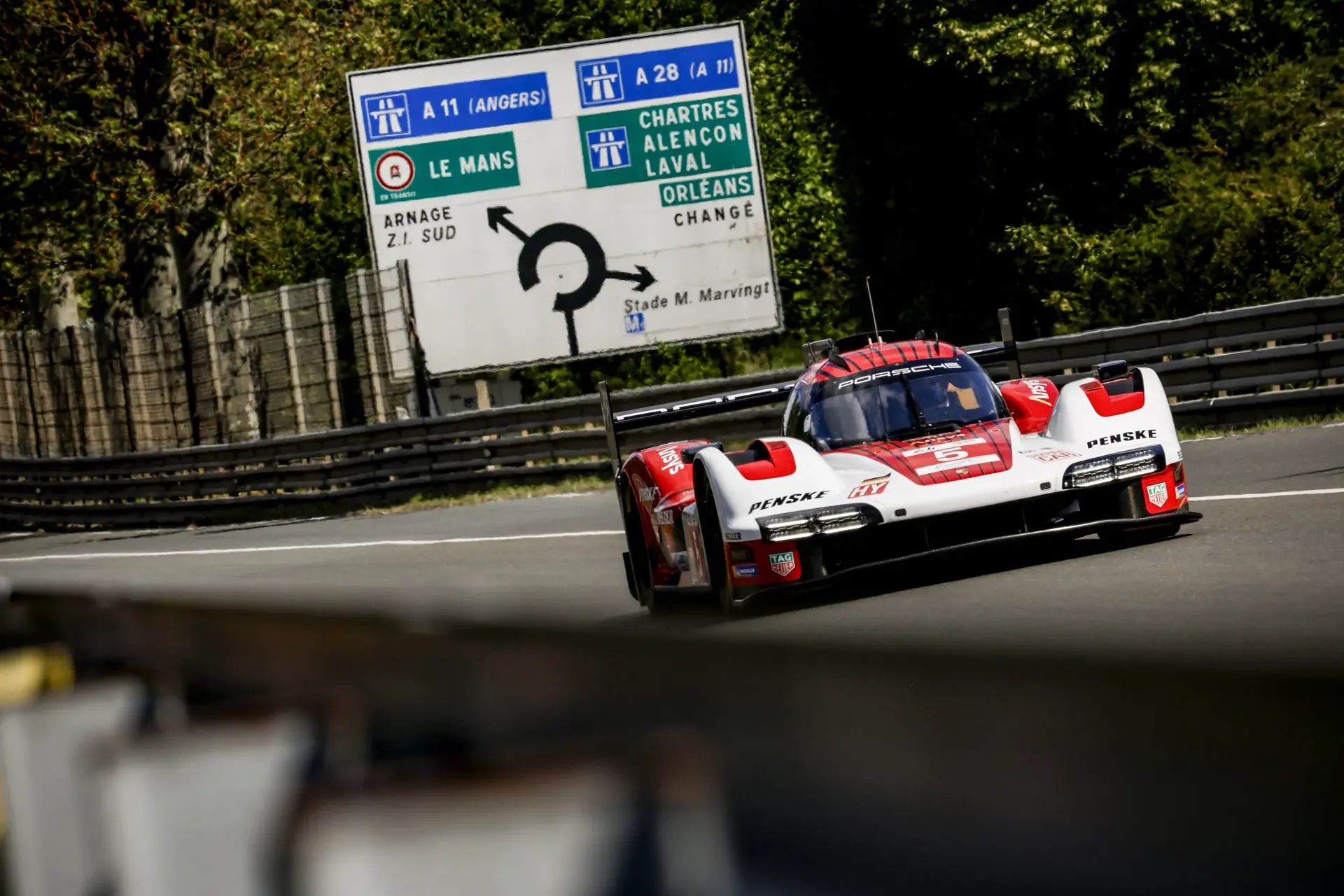En rød og hvid Porsche-racerbil kører på Le Mans-banen foran et stort fransk motorvejsskilt.