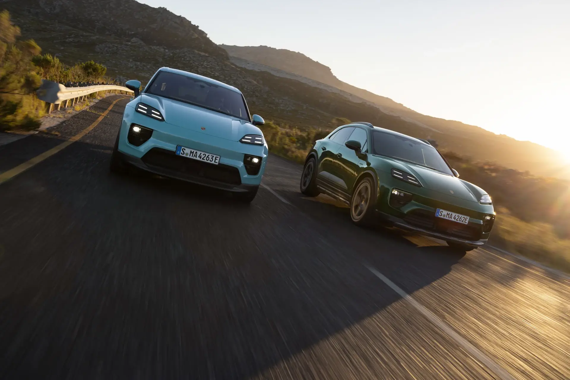 To elektriske SUV’er kører side om side på en solbeskinnet landevej gennem et bjergrigt landskab.