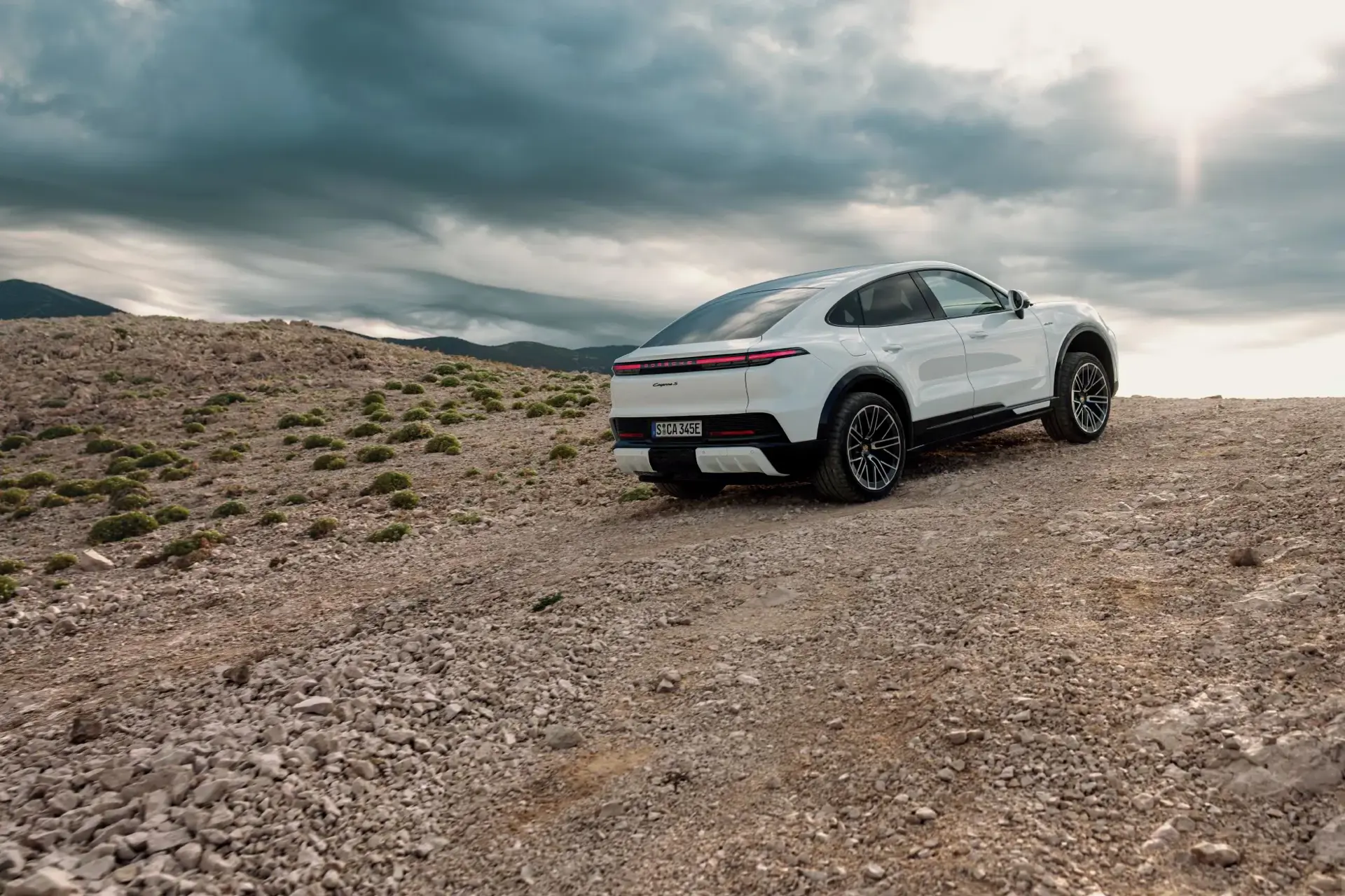 Hvid elektrisk Porsche Cayenne coupé ses bagfra, parkeret på en stenet bakke under dramatisk overskyet himmel.