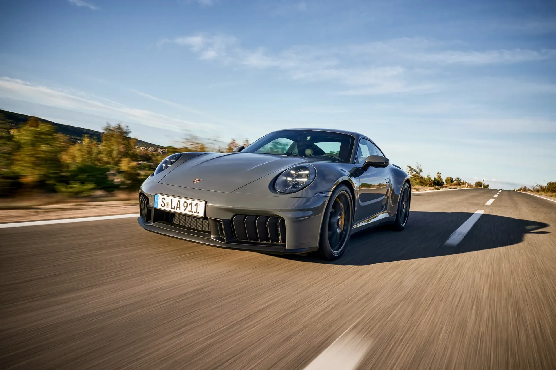 En grå sportsvogn (Porsche 911) kører hurtigt ad en øde landevej under en blå himmel.