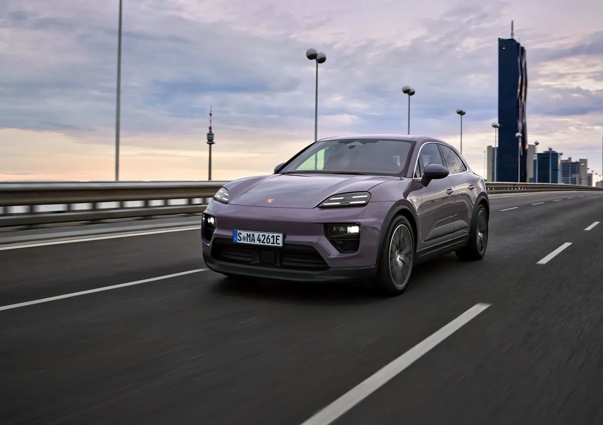 Lilla Porsche Macan kører på en næsten tom motorvej med en moderne byhorisont i baggrunden ved solnedgang.