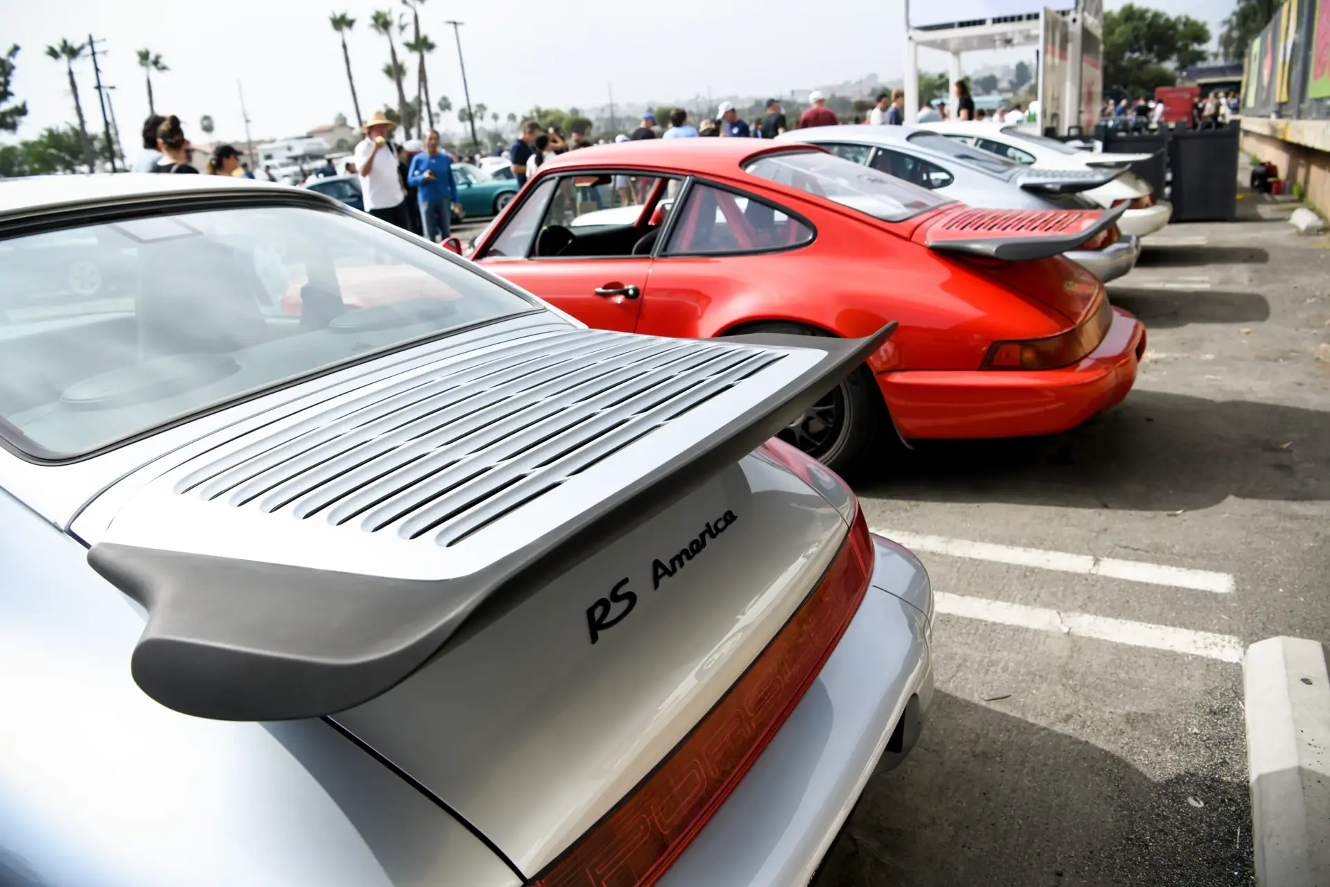 Række af klassiske Porsche 911 sportsvogne, heriblandt en sølvgrå RS America, parkeret ved et biltræf udendørs.