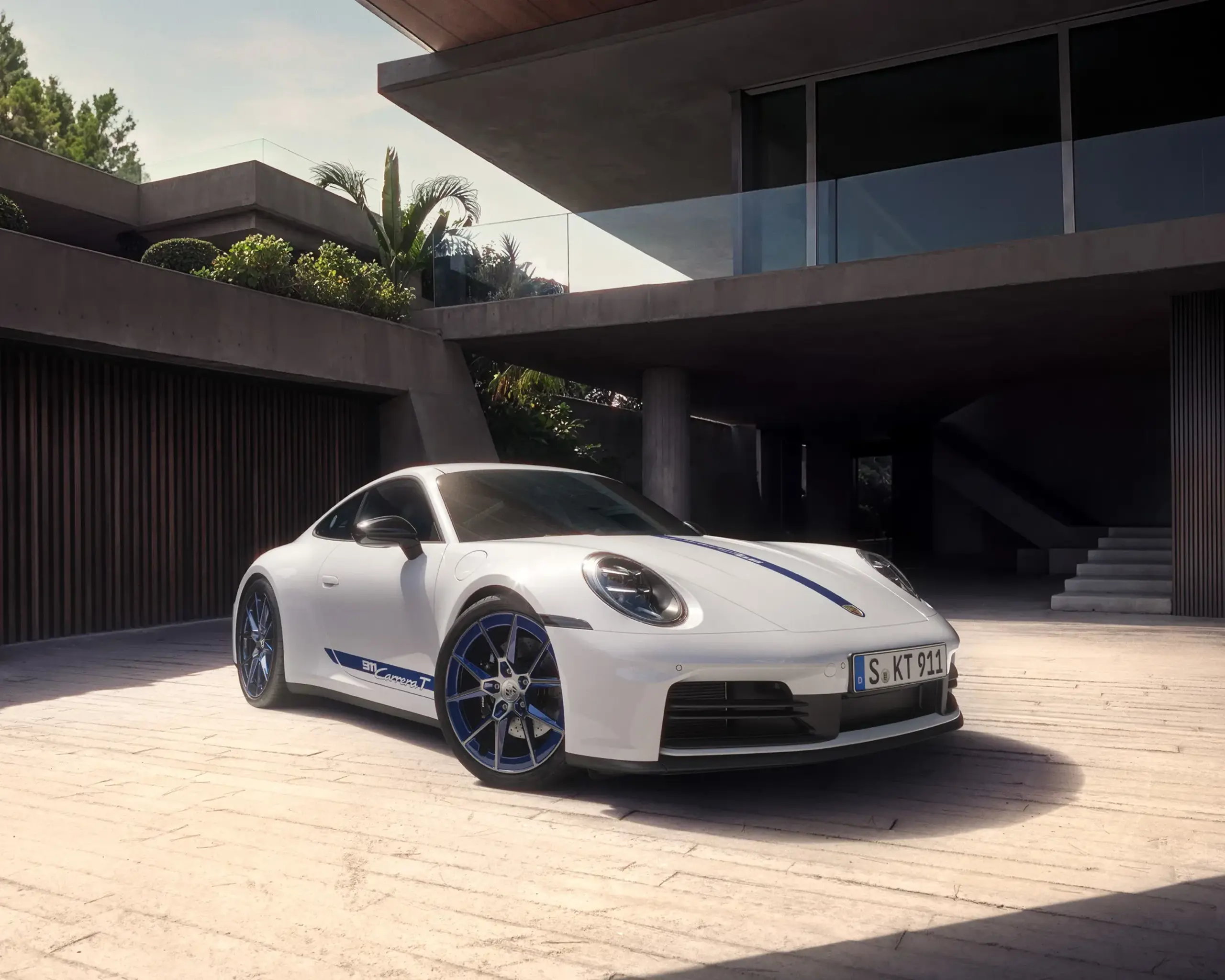 En hvid Porsche 911 Carrera T holder parkeret i sollys på en moderne, træomkranset betonvej.