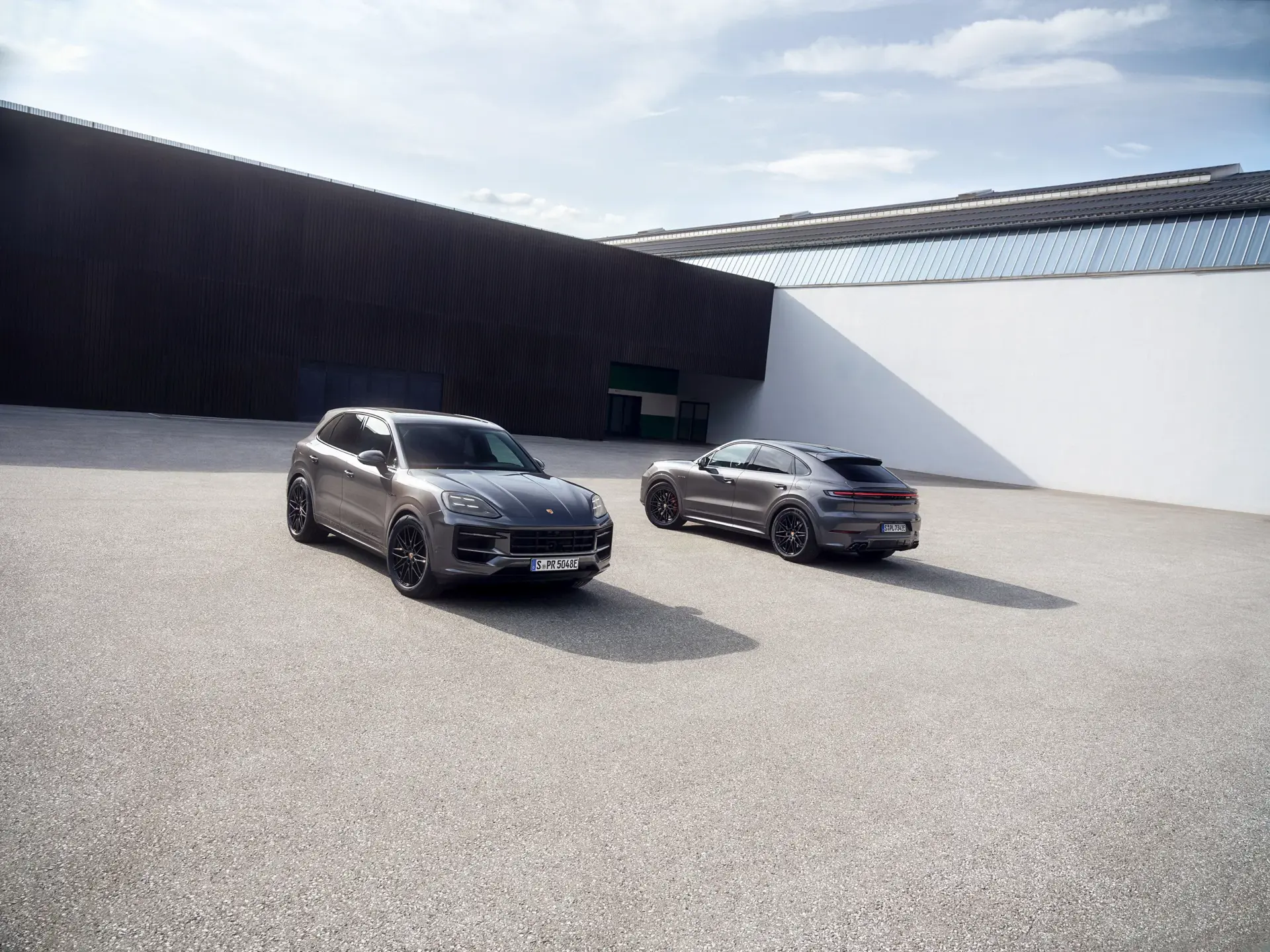 To grå Porsche Cayenne SUV-modeller holder parkeret på en stor, tom plads foran en moderne industribygning.