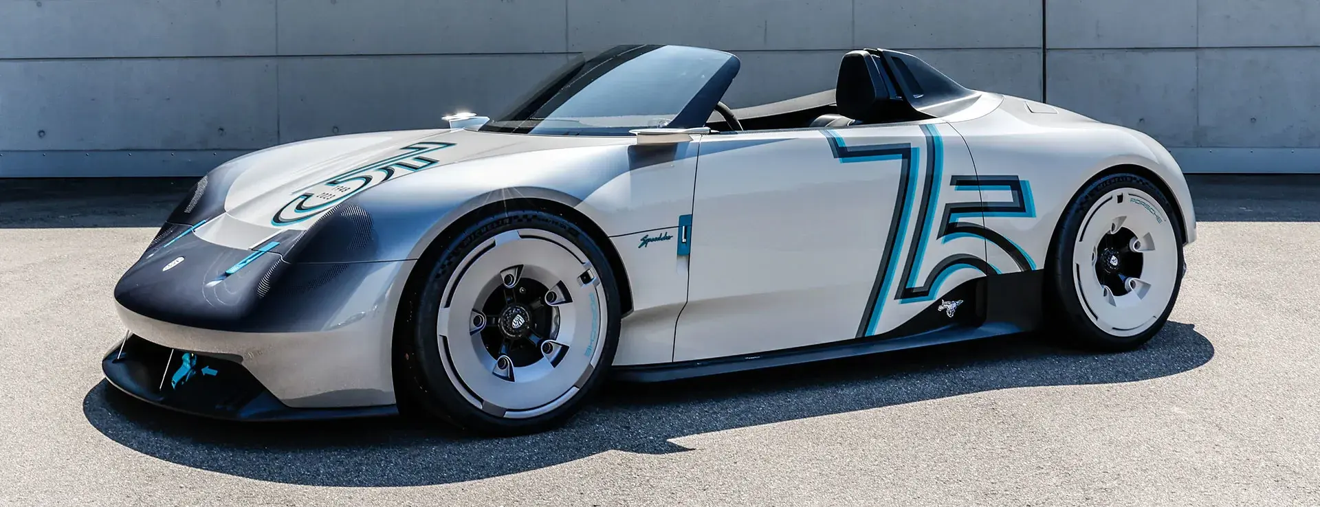 En sølvfarvet Porsche Vision 357 Speedster sportsvogn uden tag holder parkeret foran en grå betonmur.