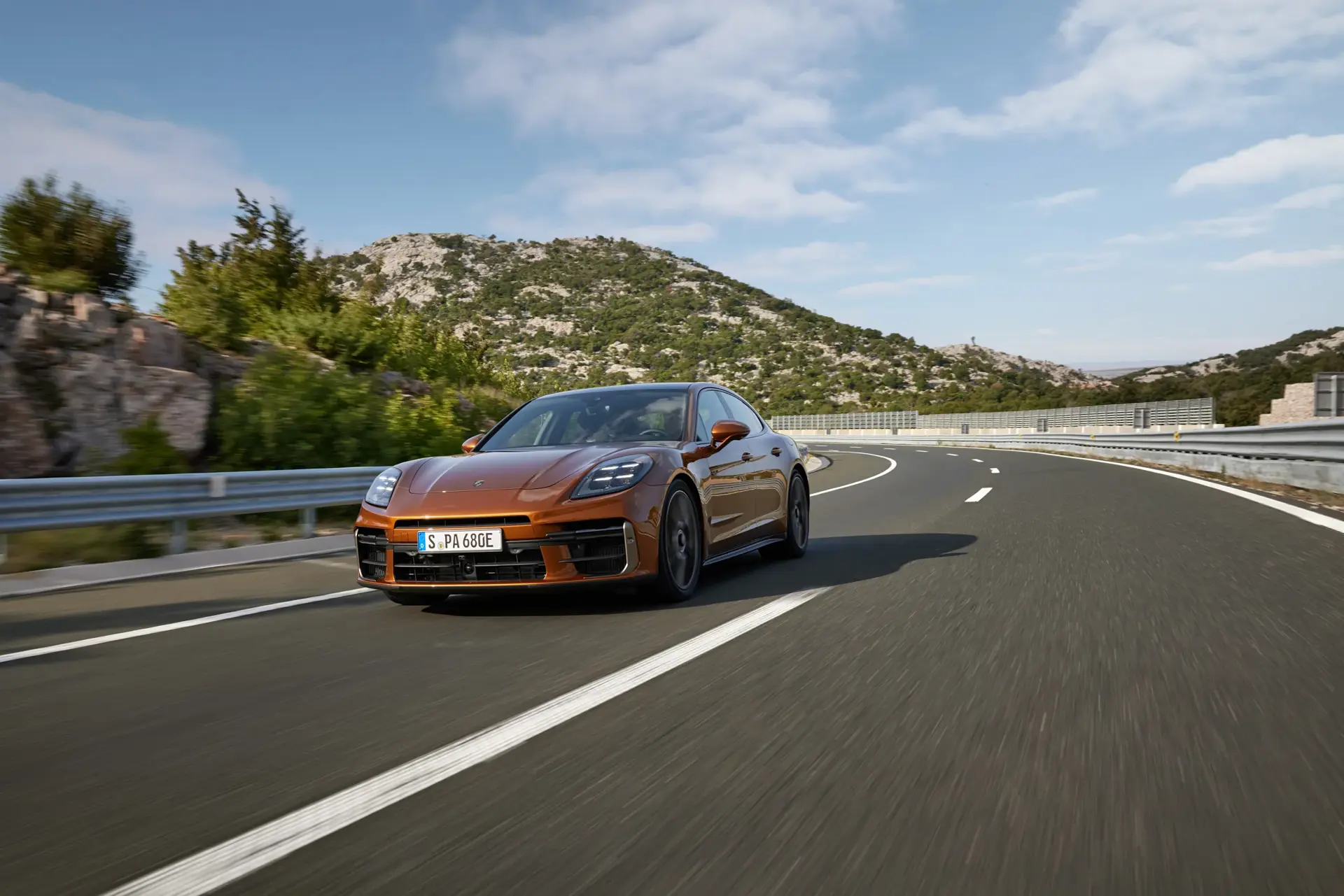 En bronzefarvet Porsche Panamera kører på en tom, snoet motorvej gennem et bjergrigt landskab under blå himmel.