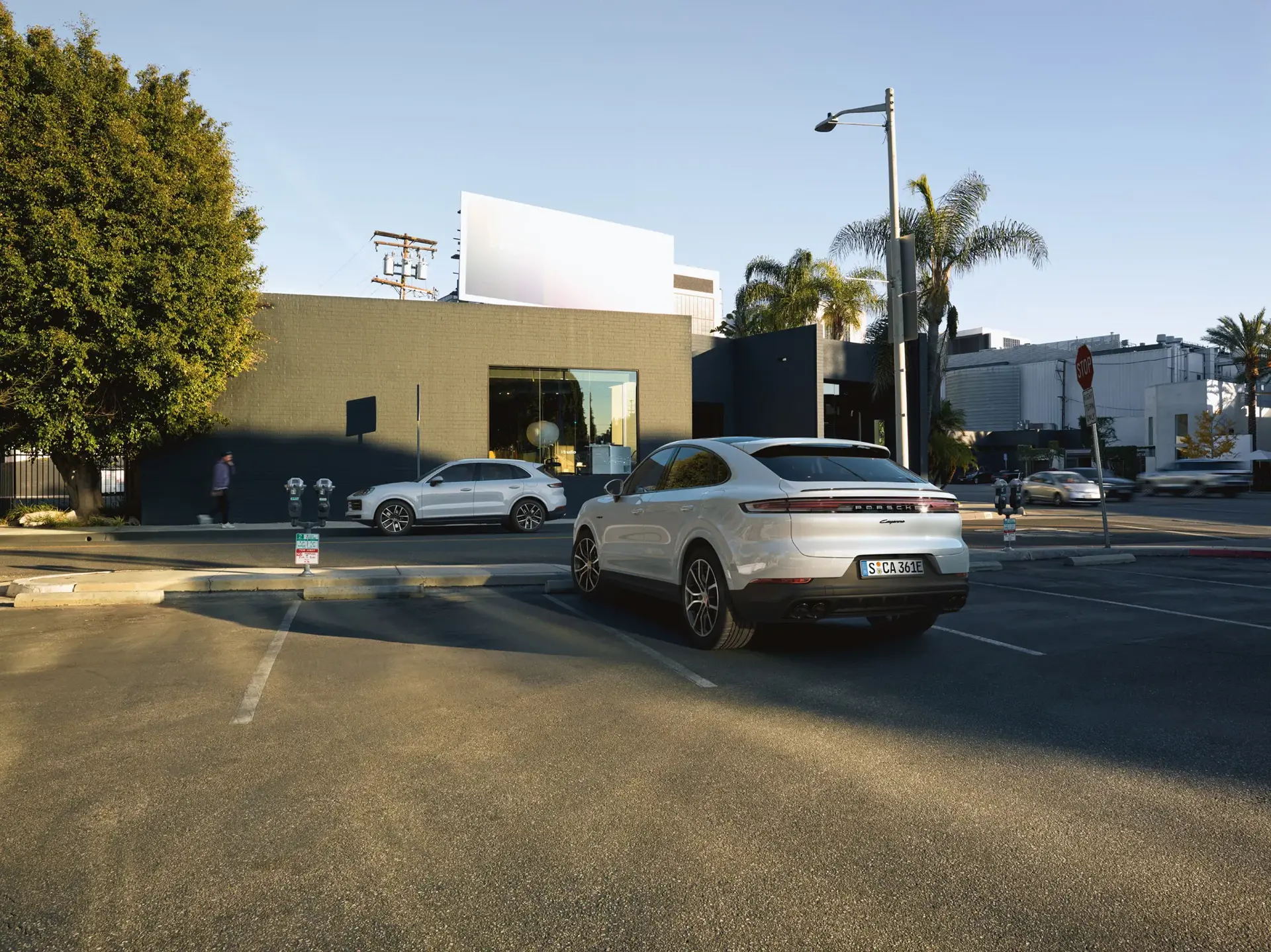 En sølvfarvet Porsche SUV holder parkeret på en solrig gade foran en moderne bygning med palmer i baggrunden.